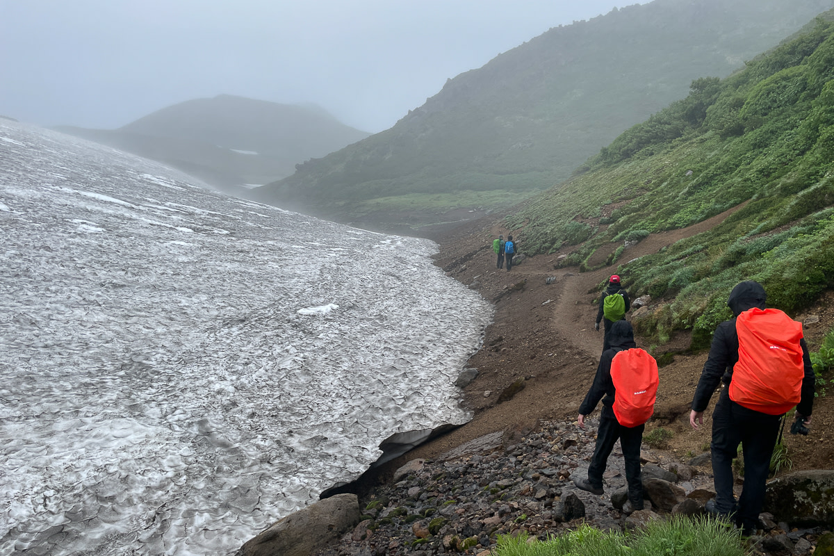 A group of hikers walk past a large patch of snow amongst misty mountains