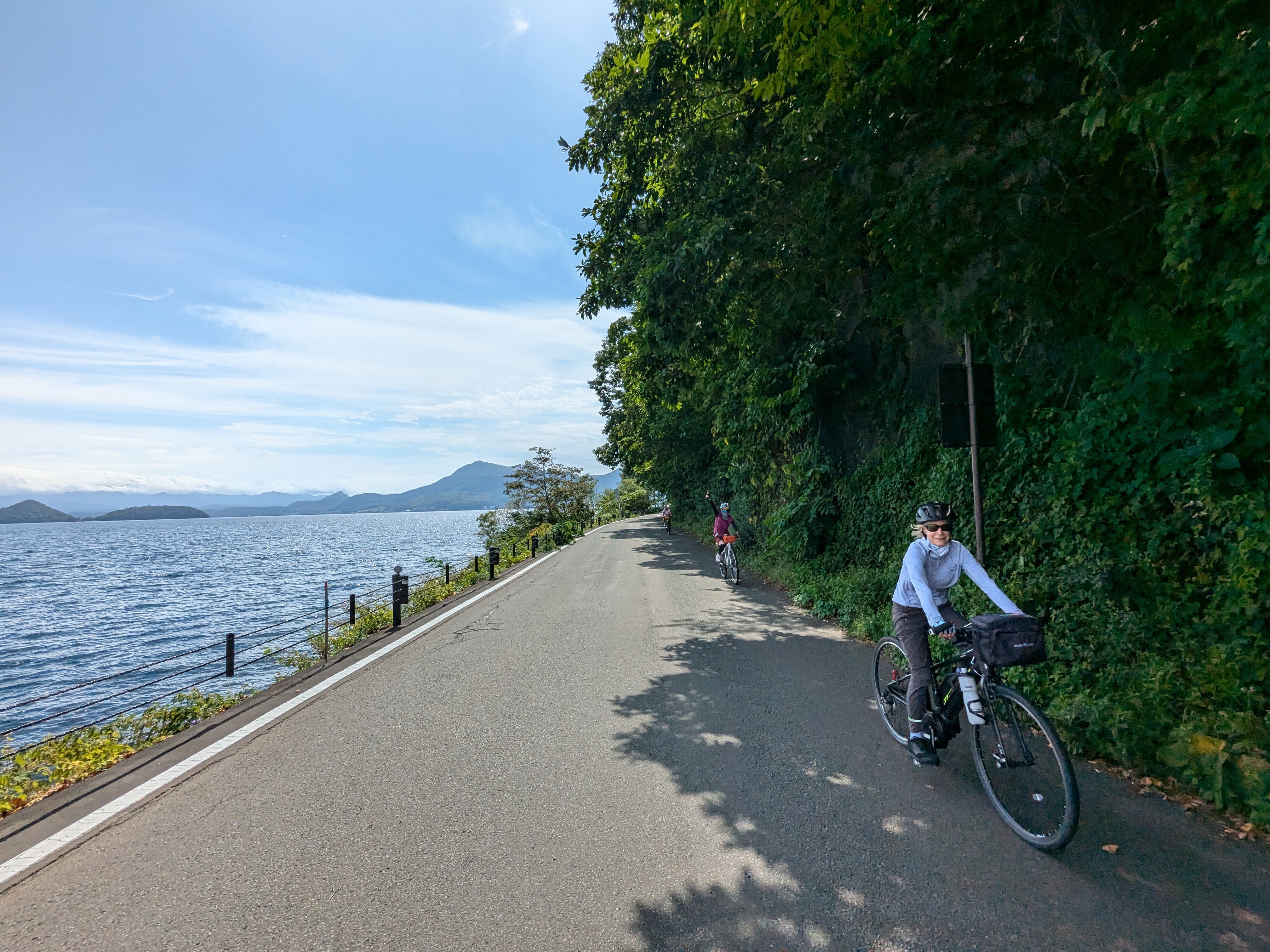 Two cyclists cycle on a road alongside Lake Toya, Hokkaido. It is a very sunny day. The woman at the front of the group smiles at the camera as she cycles past.