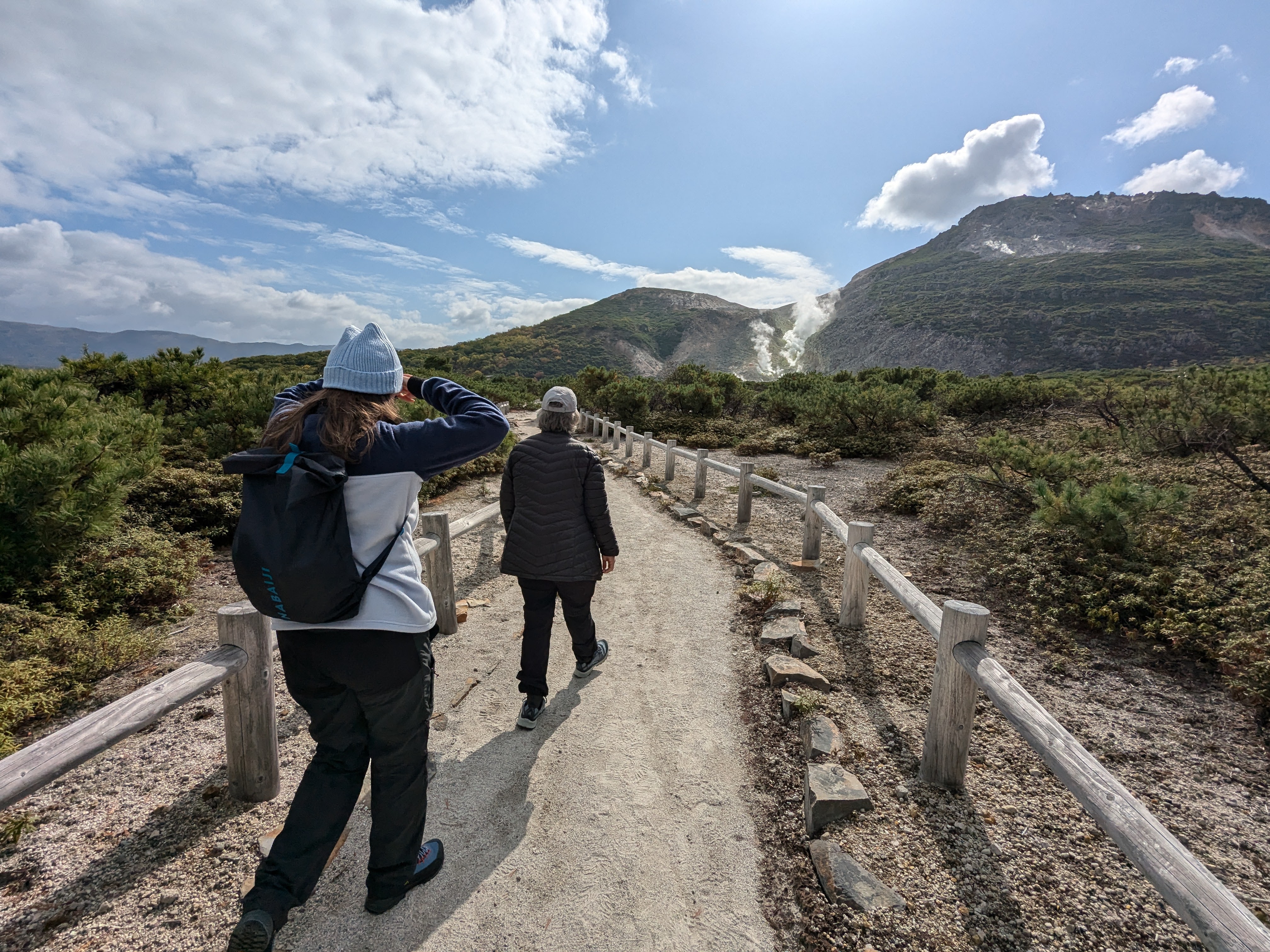 Two walkers approach Mt. Io via a fenced walking path. Mt. Io is in the distance. It is an active volcano with steam rising from its vents.