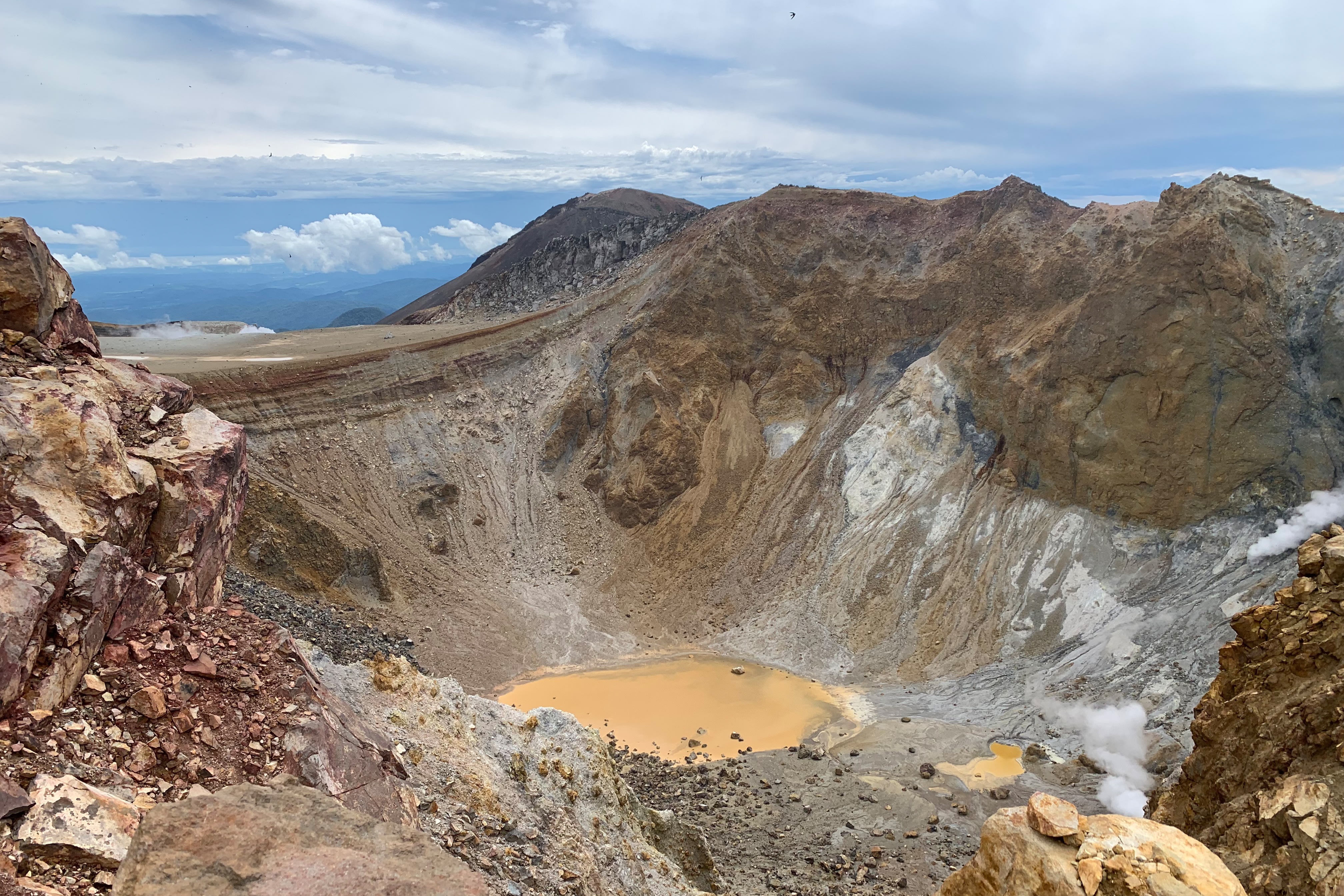 A dramatic view of a volcanic crater on Mt Meakan. There is a brown lake deep in the bottom of the crater, while steam billows up from vents.
