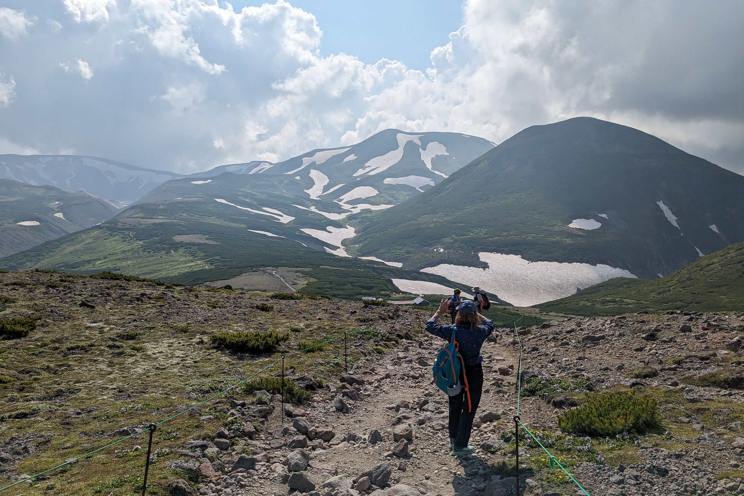 A hiker takes a photo of a cloudy sky and snow lingering on mountains along the Asahidake-Kurodake Traverse in Hokkaido.