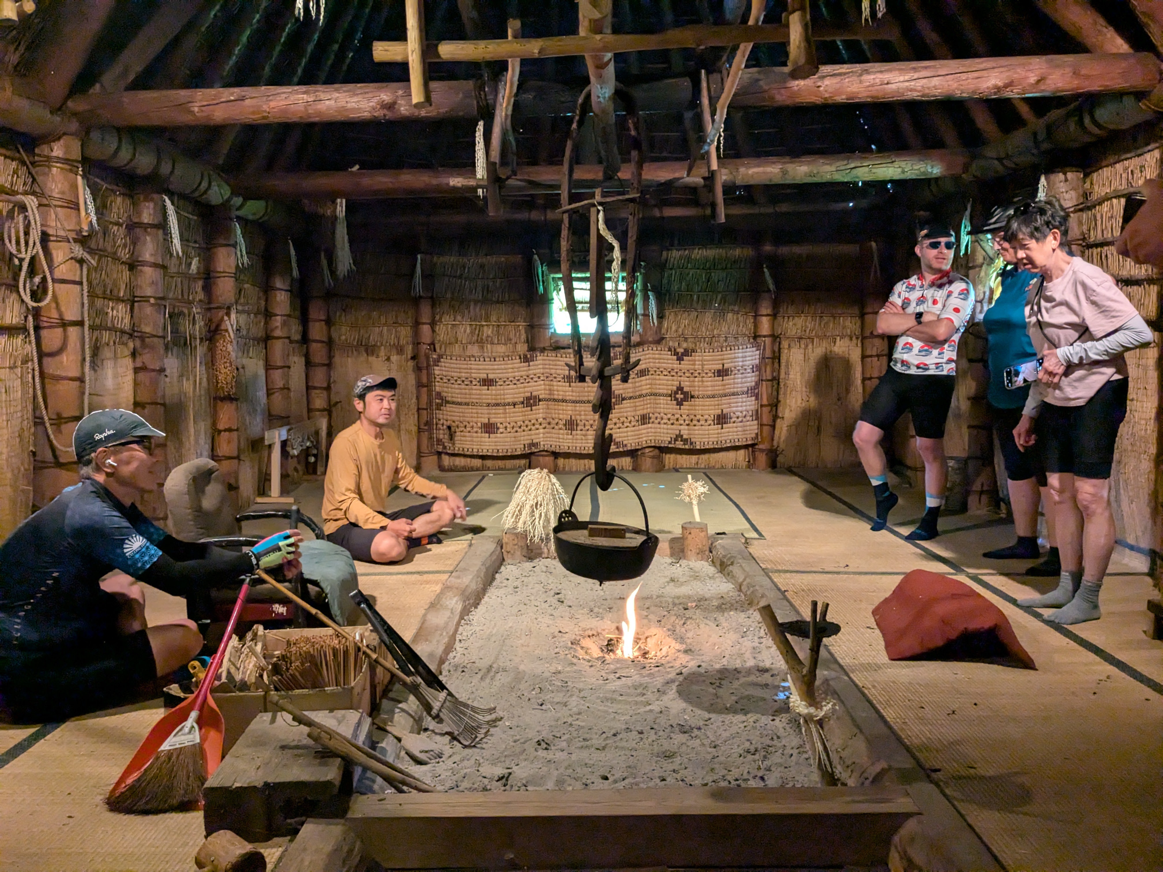 A group of cyclists taking a break inside a "cise", a traditional Ainu dwelling. In the middle of the room is a Japanese hearth with a suspended pot being warmed by an open flame.