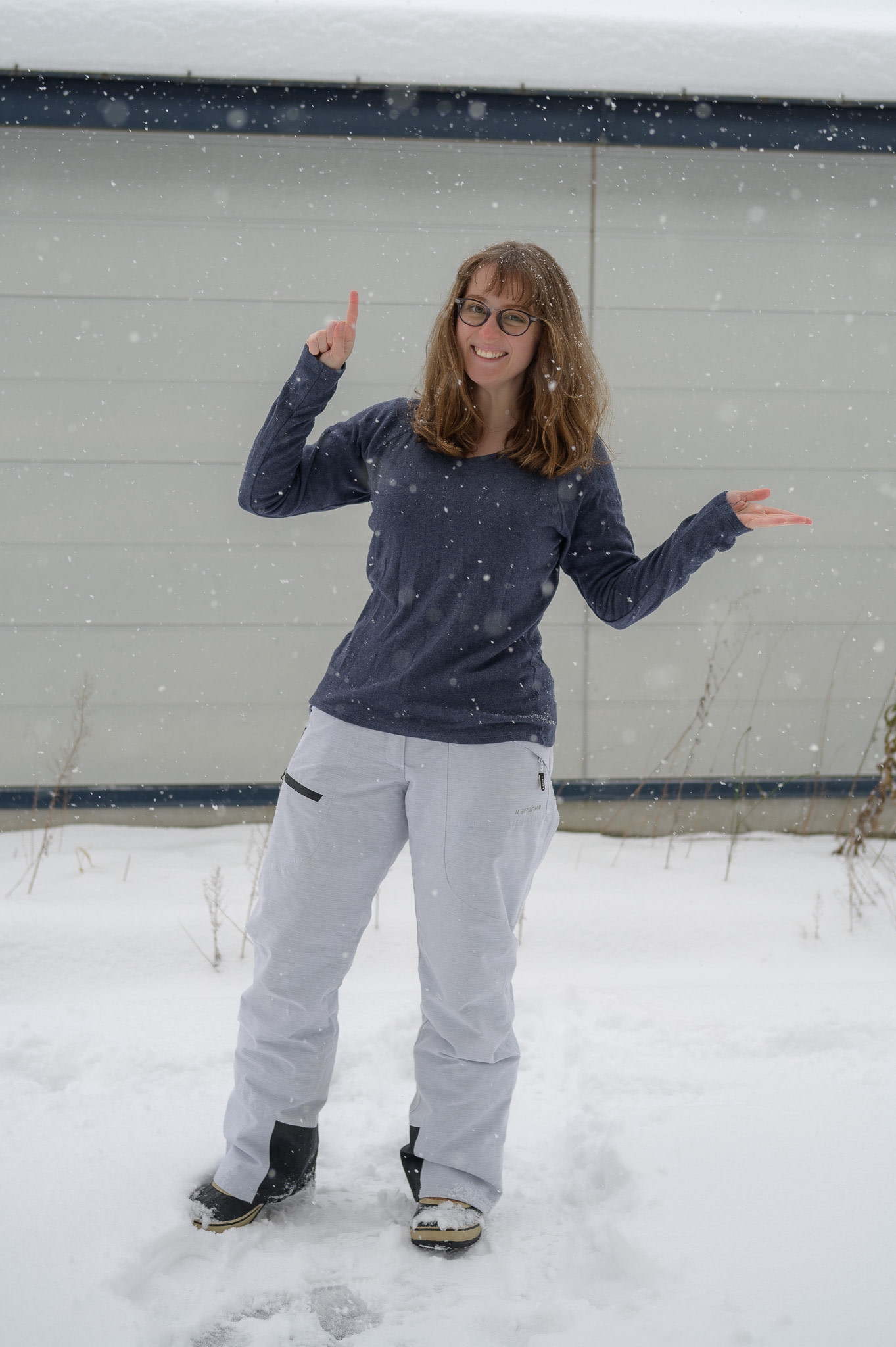A girl wearing glasses stands in the snow wearing ski pants and a fleece top.