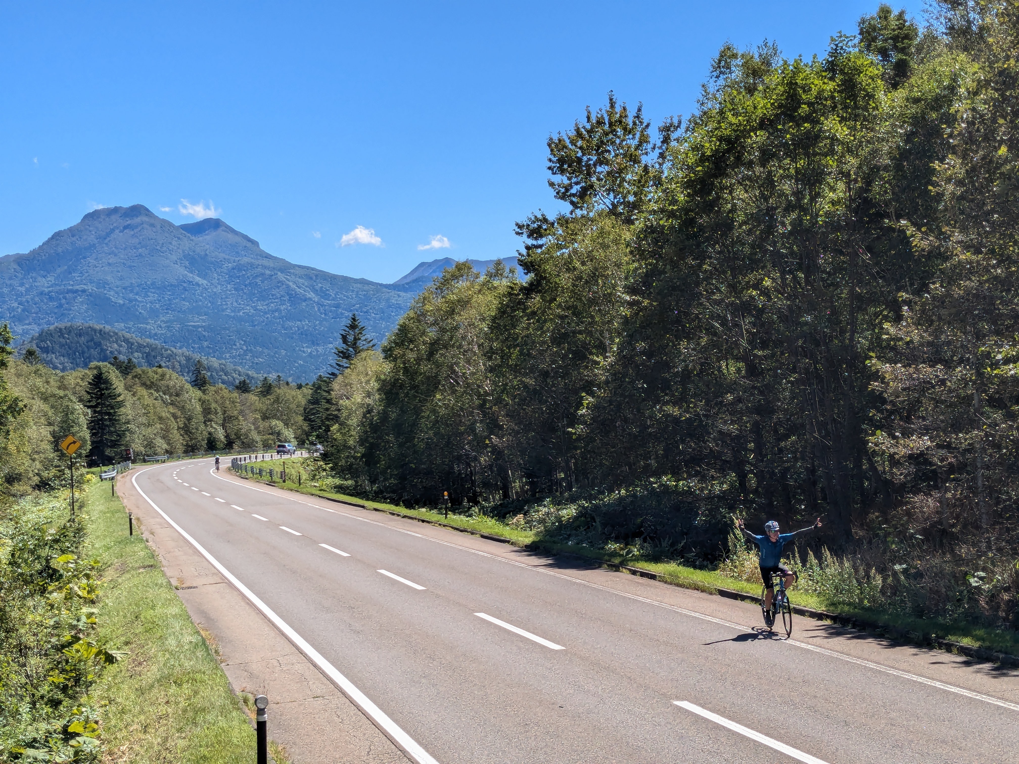 A high angle view of a cyclist in a mountain pass. They have their arms raised and are smiling at the camera. It is a beautifully sunny day and a mountain is visible in the background.