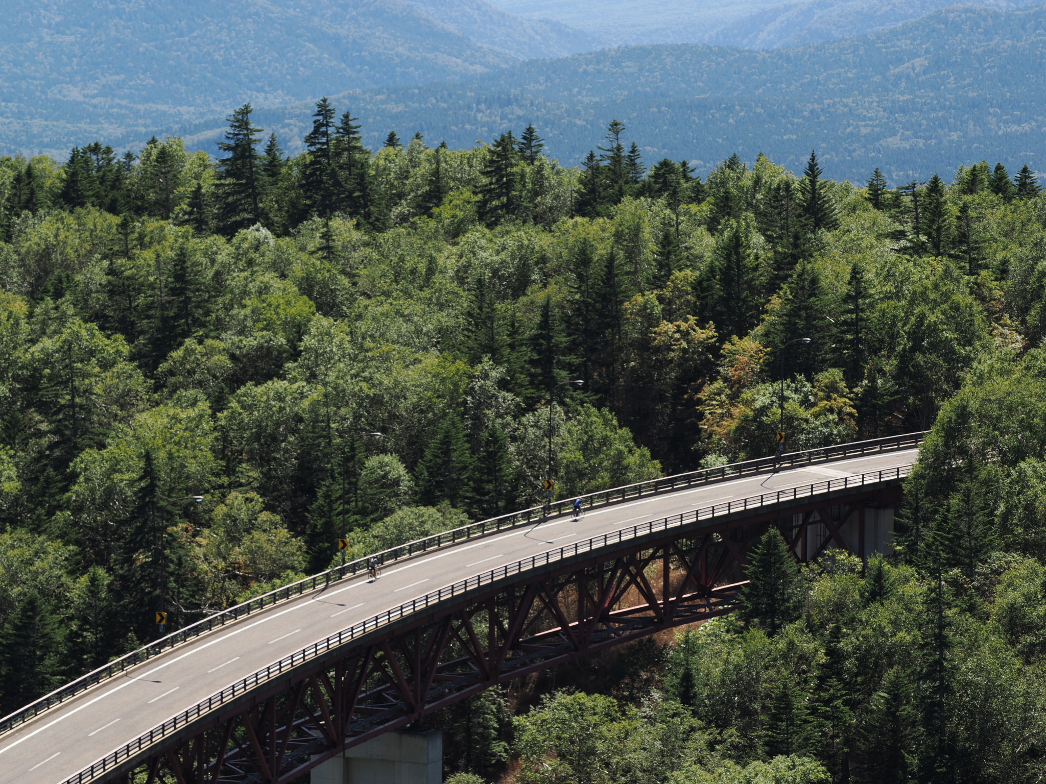 A zoomed view of a large bridge spanning a forested, mountain valley. Two cyclists are just visible crossing the bridge.