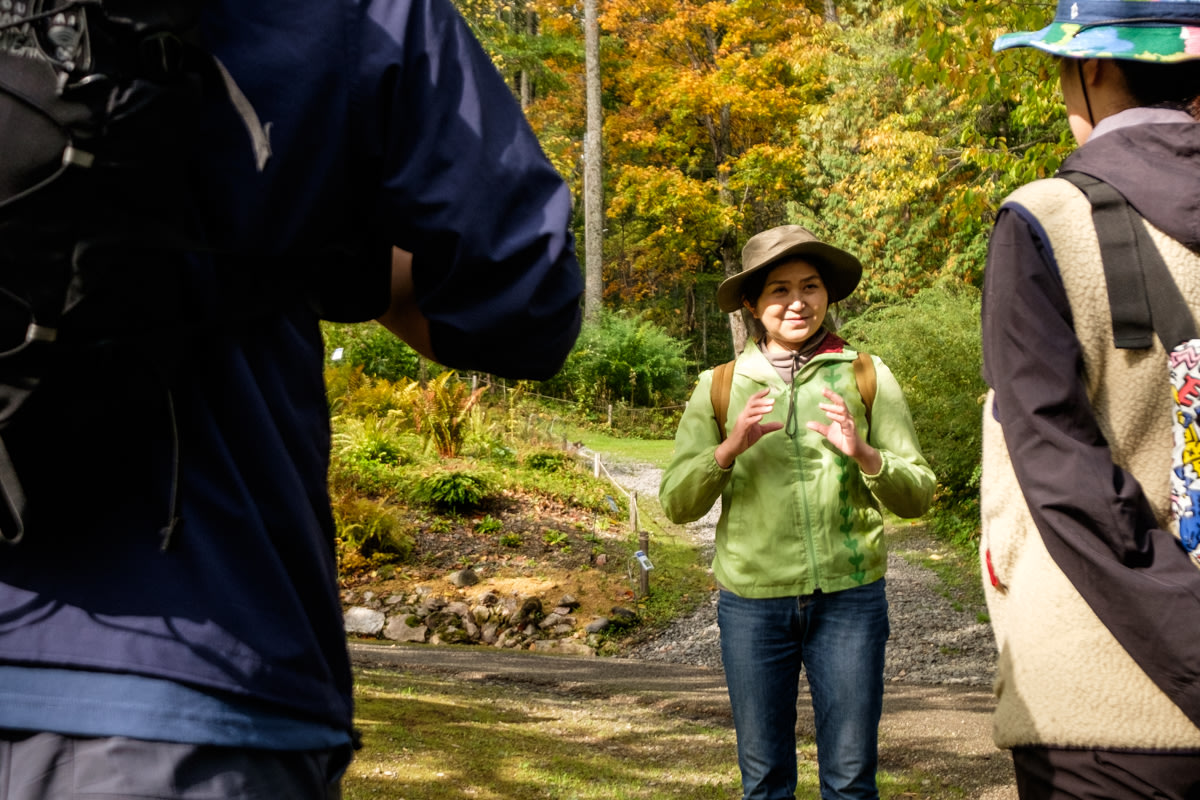 An Ainu guide in modern clothing stands in front of the entrance to a mountain trail, explaining something to two people in the foreground.