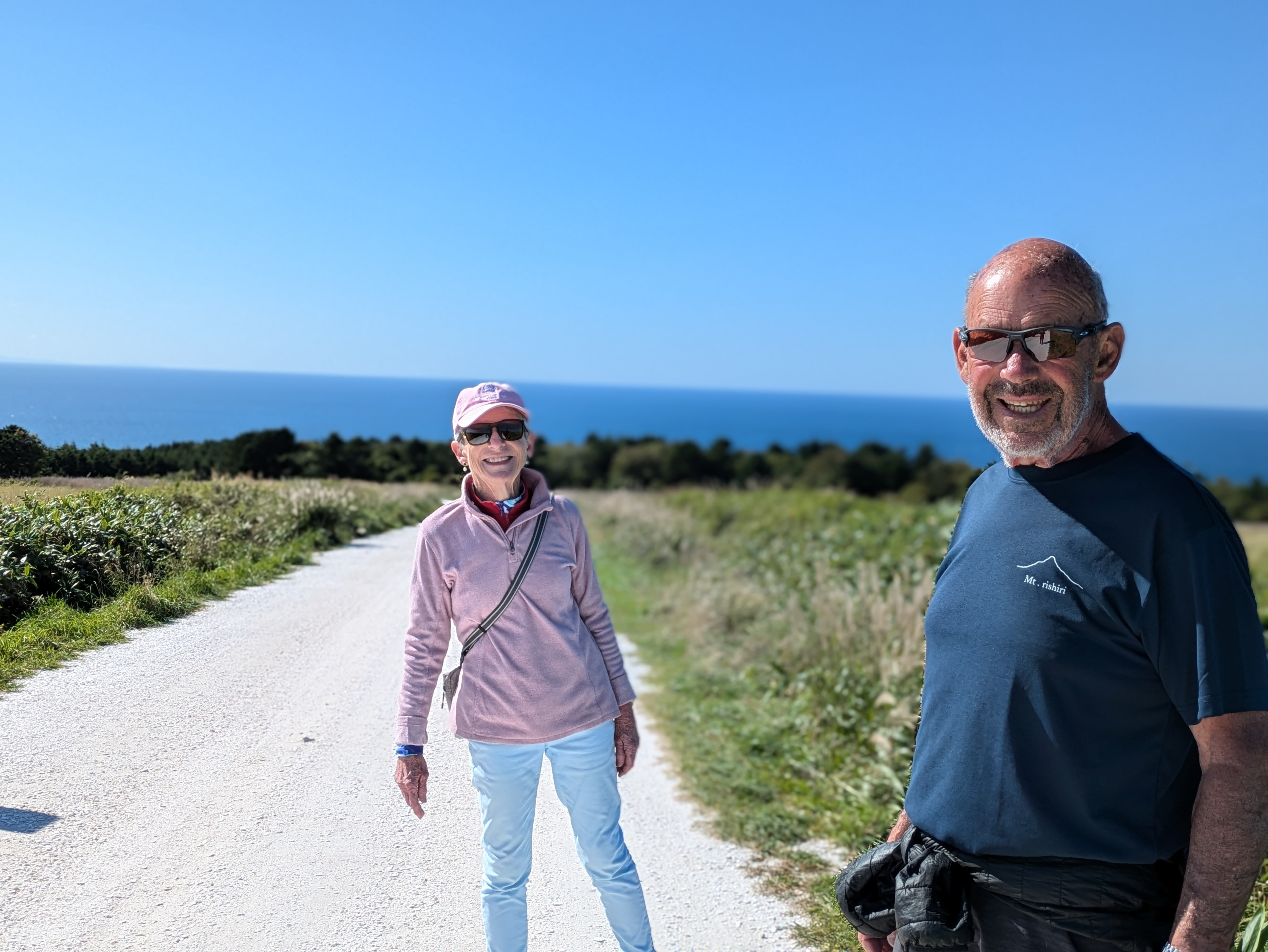 A man and a woman smile at the camera, stood on the White Shell Path, Wakkanai, Hokkaido. The path is made from crushed white seashells, hence its name. It is a very sunny day and the ocean is visible in the distance.