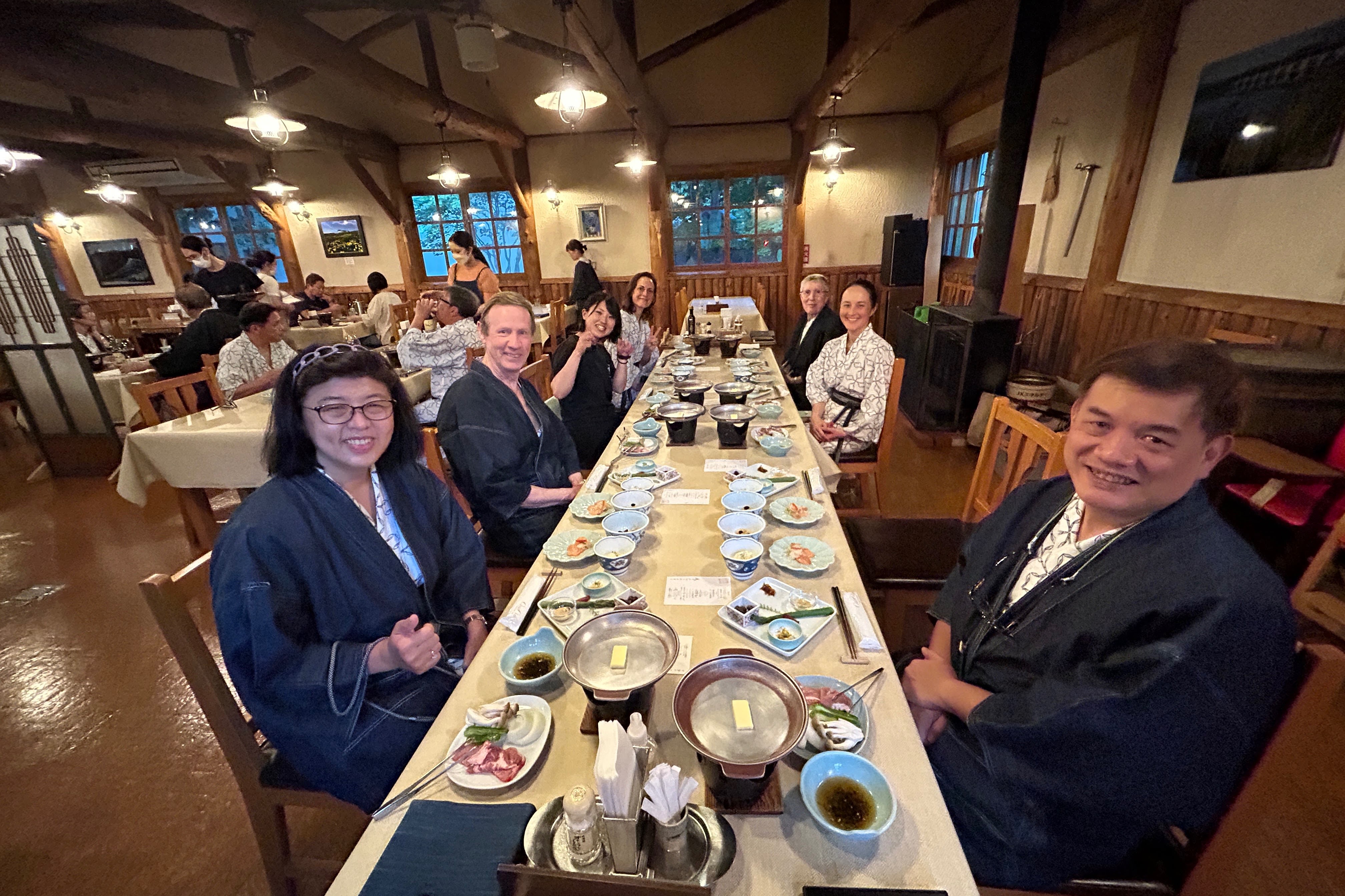 A group of people sit at a long table laden down with food and smile at the camera. They are all wearing yukata, traditional Japanese bath robes.