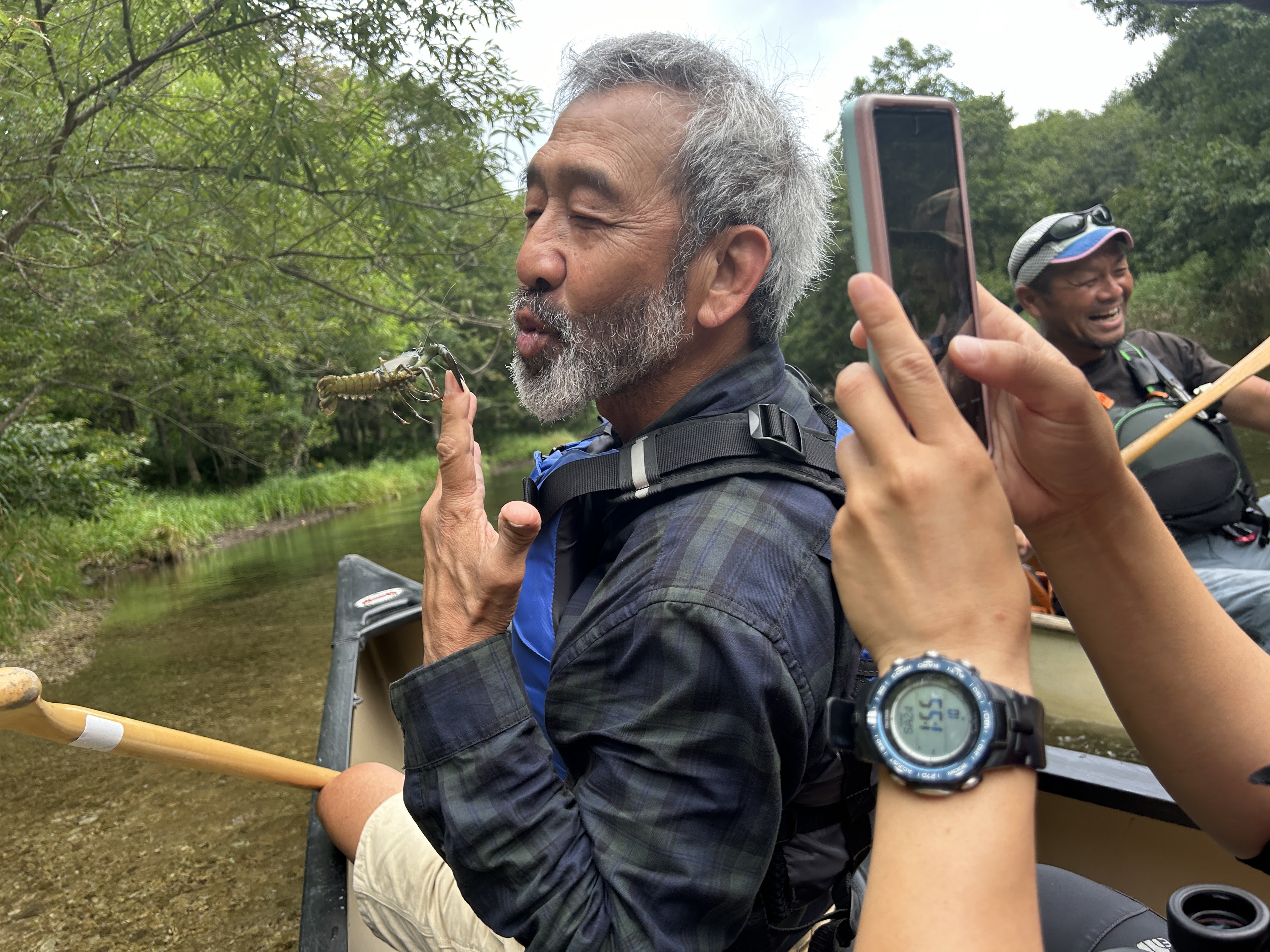 A man jokes around with a crayfish pinching his finger! He is holding it up to his mouth as if to kiss it. Someone in the foreground has raised their smartphone and is taking a photo of the scene.