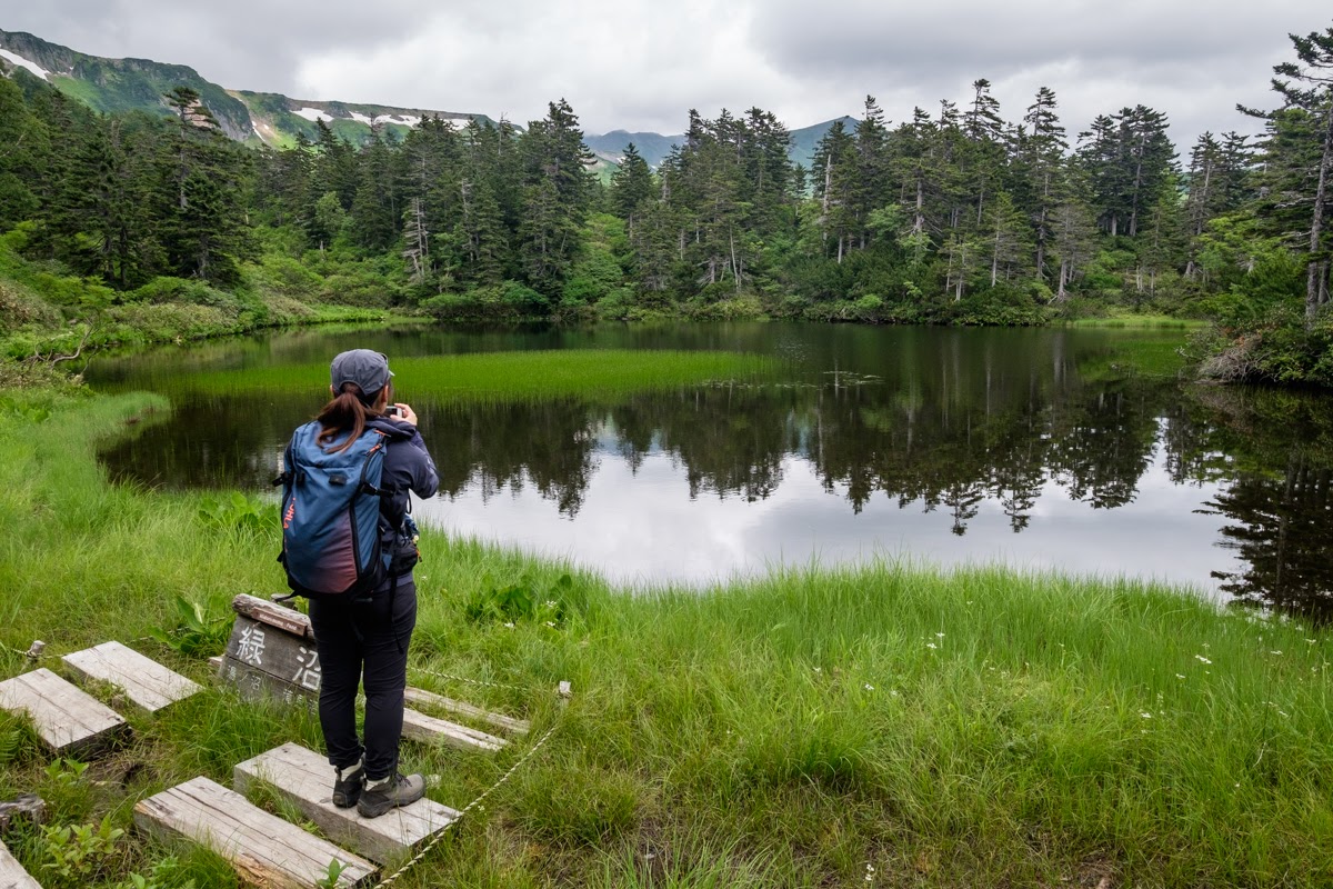 A hike stops to take a photo of Midori pond at Daisetsuzan Kogen. In the distance snow clings to the ridgeline.