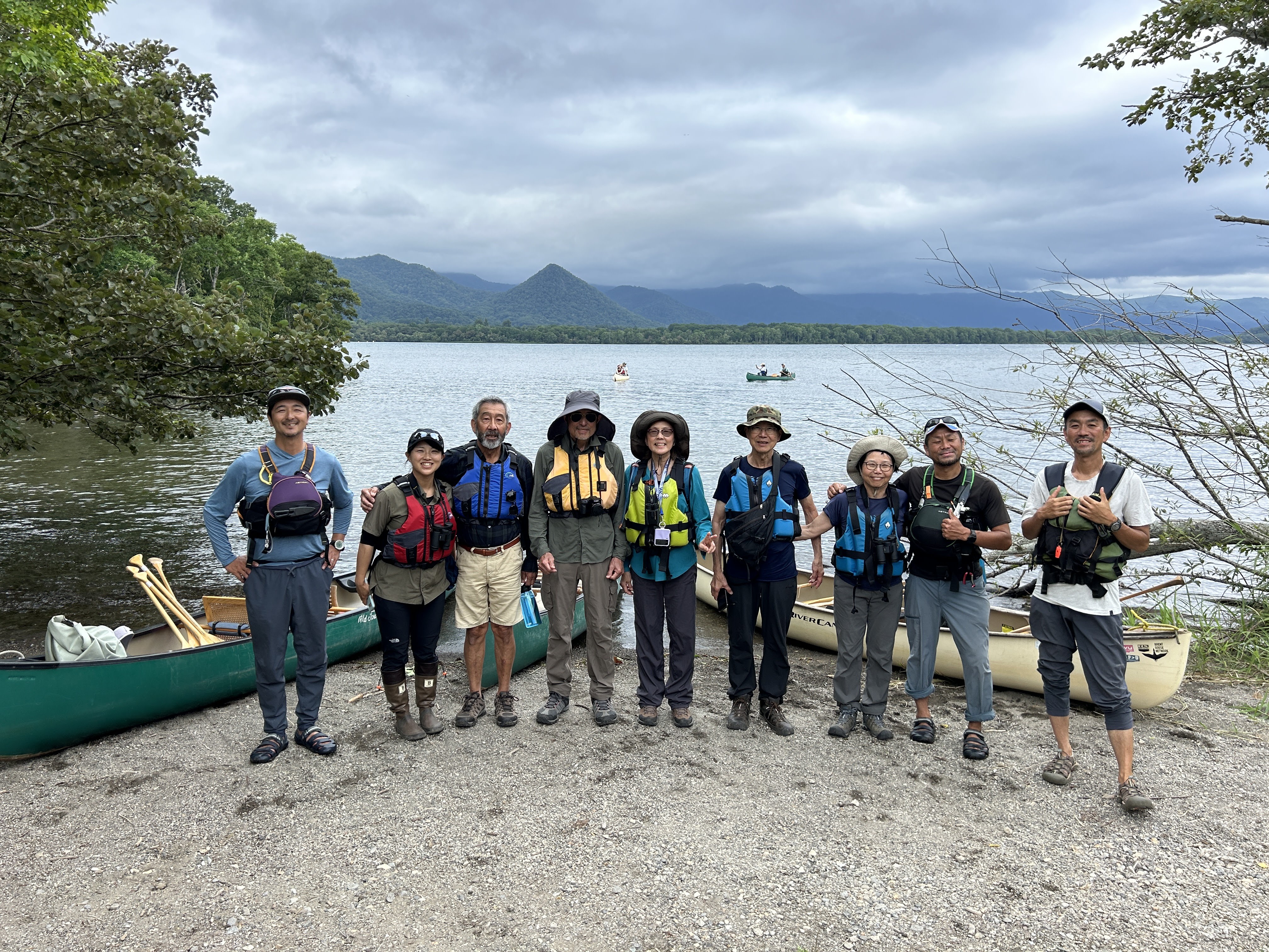 A group of people stand in a line on a lakeshore, canoes behind them. They are all smiling at the camera and wearing life jackets, ready to go canoeing.