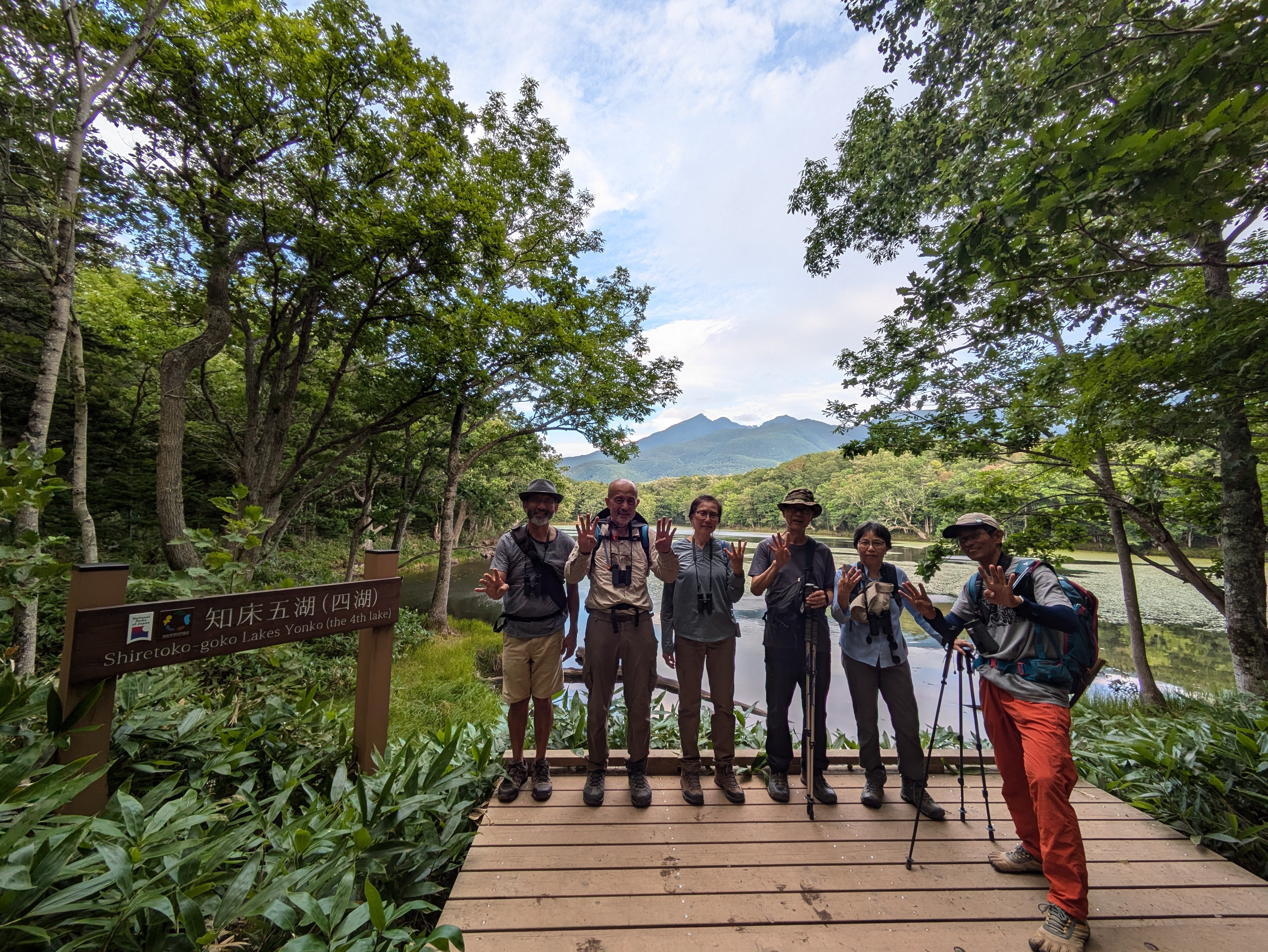 A group of six people stand on a boardwalk in a forested lakeside. There is a lake behind them and a mountain in the distance. A sign to their left reads "Shiretoko Goko Lakes Yonko (The Fourth Lake)". Everyone in the group is holding up four fingers.