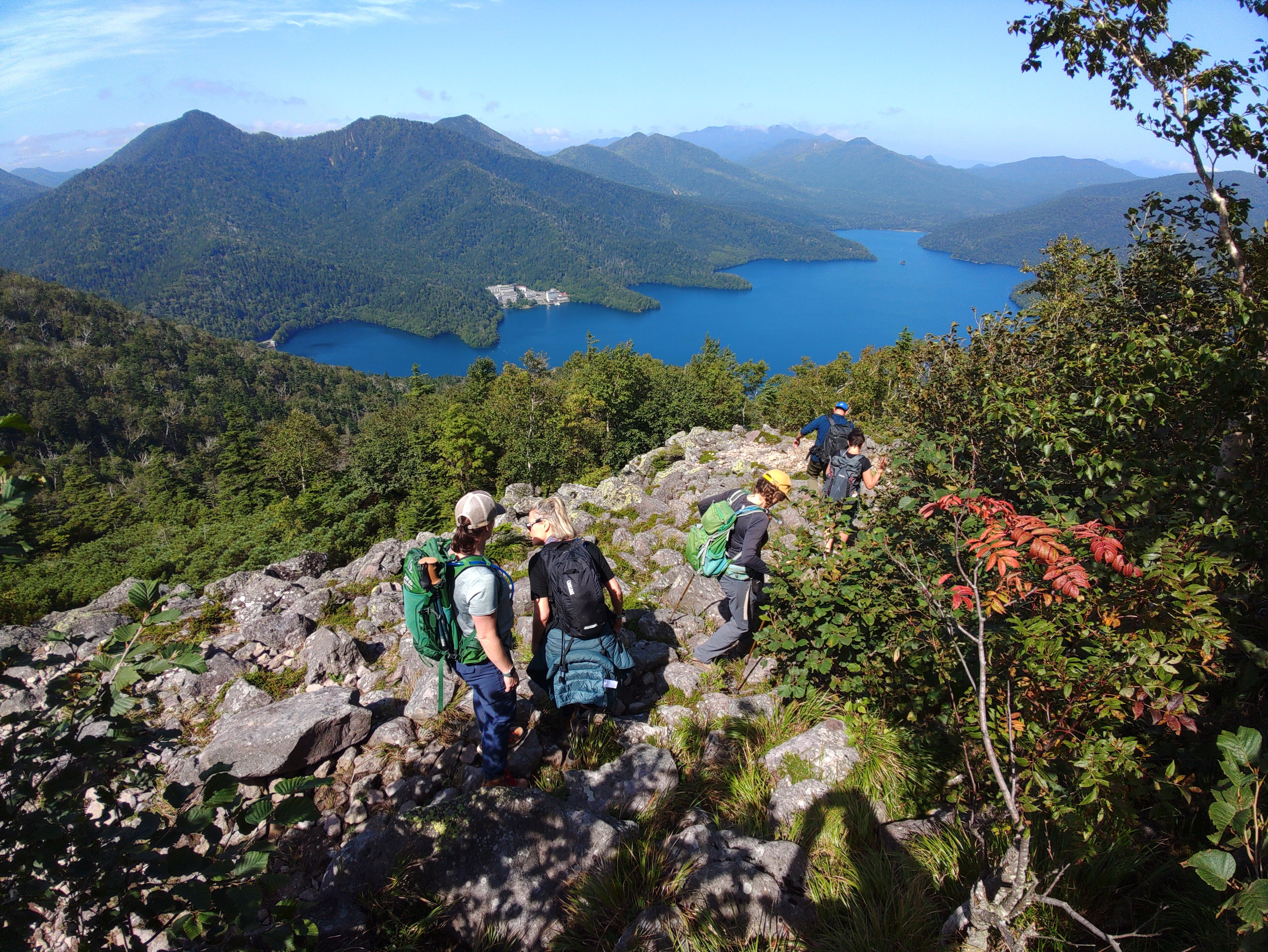 Hikers begin to descend from the rocky summit of Mt. Hakuunzan in the Daisetsuzan National Park. It is a clear day and Lake Shikaribetsu stretches out below the open summit area.