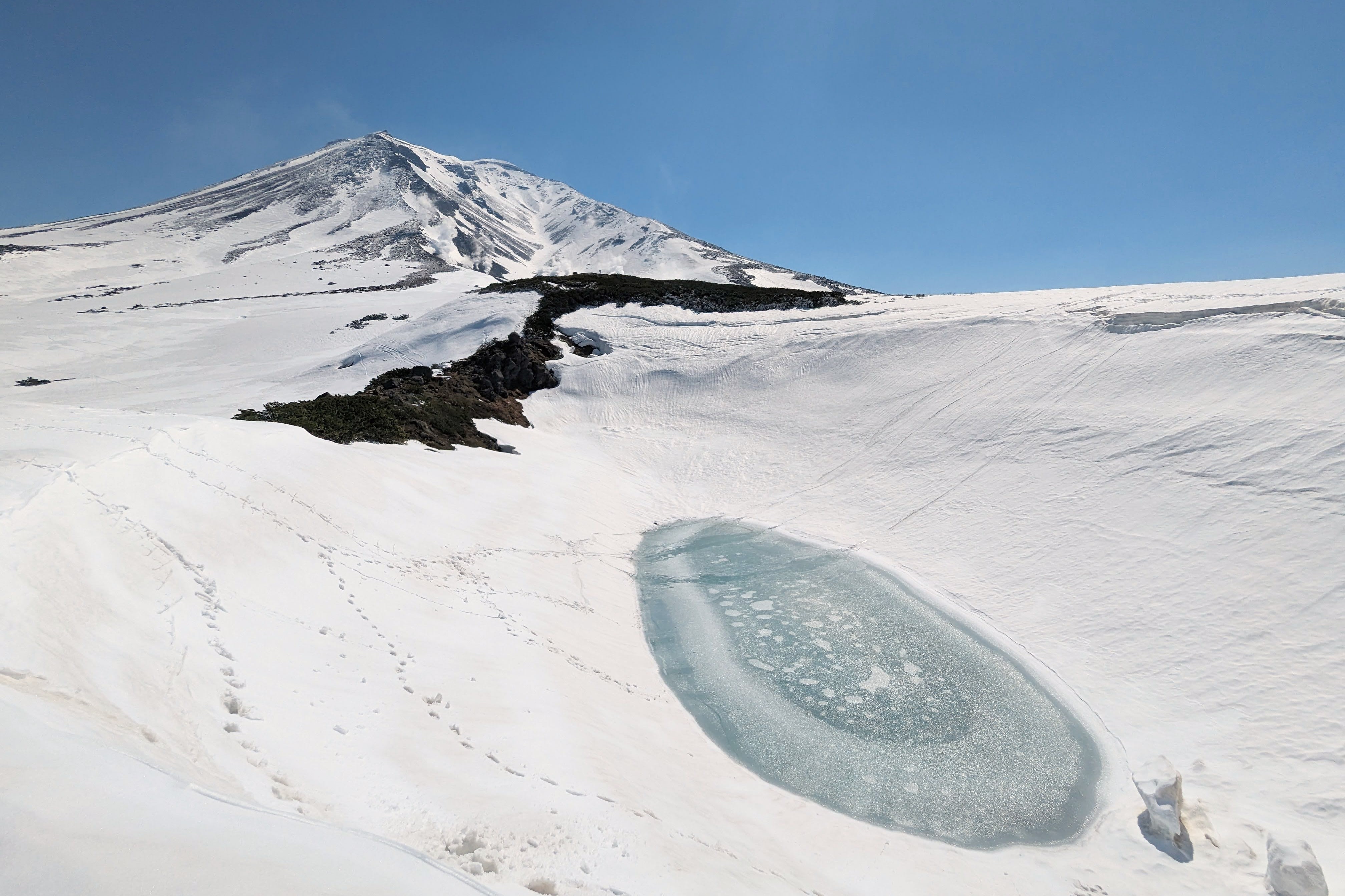 A frozen pond is surrounded by snow. Mt. Asahidake stands above the pond mostly covered in snow bar some rocky ridge lines. Steam can be seen rising from fumeroles in the distance. The sky is blue and cloudless. In the foreground a number of snowboard tracks can be seen crossing the pond.