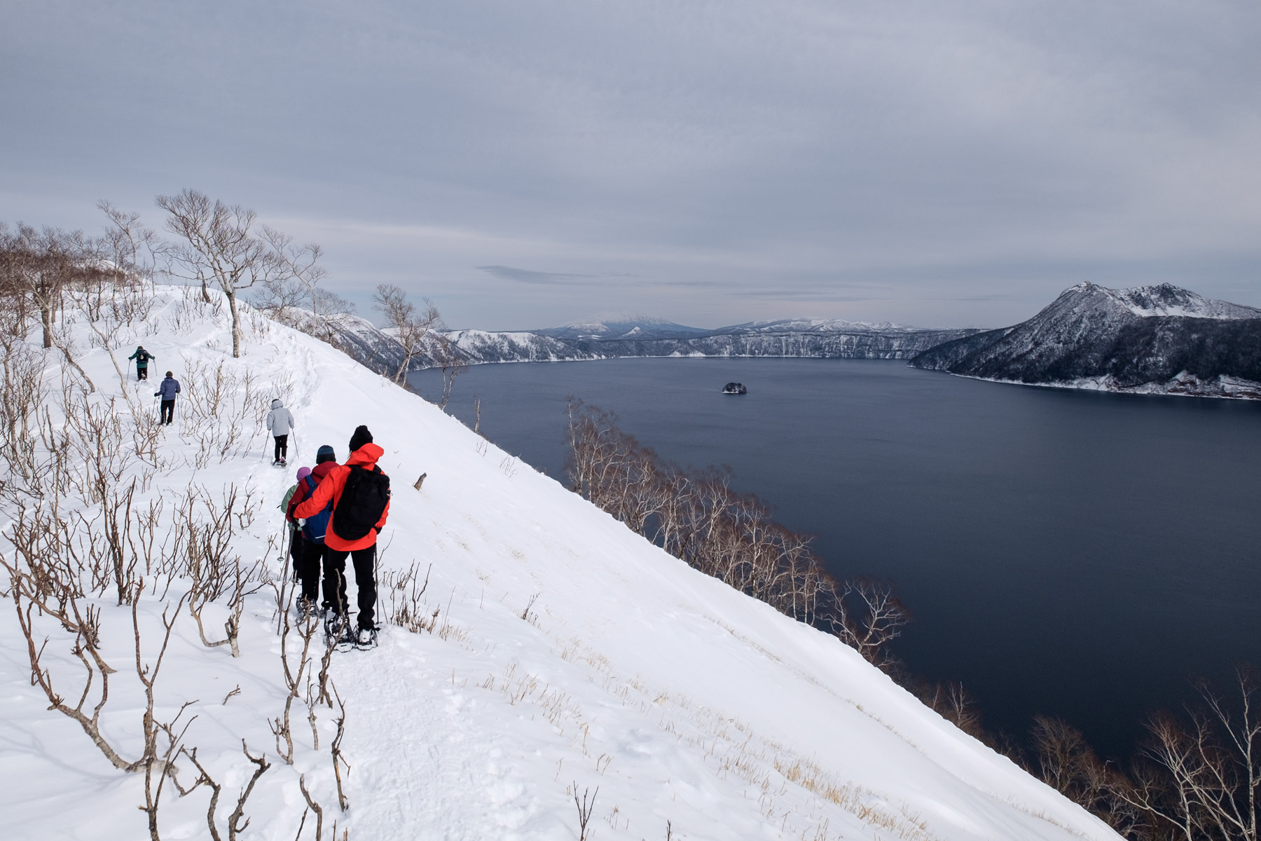A snowshoeing group walks amongst birch trees along the edge of the Lake Mashu caldera. Hundreds of metres below the lake is still and a steel grey under an overcast sky. In the distance Mt Mashu can be seen rising above the opposite side of the lake.