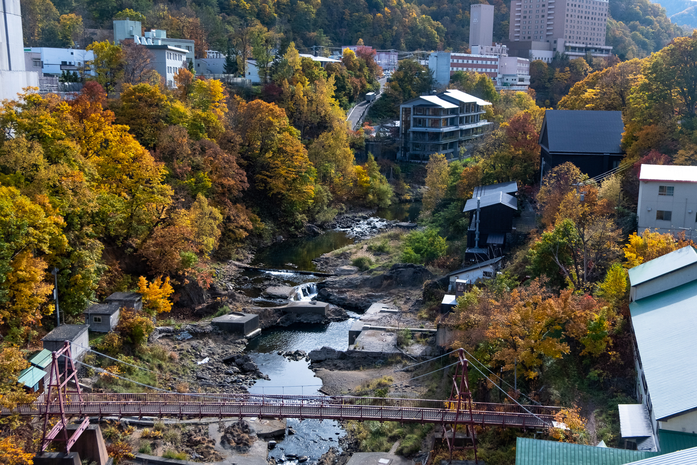 An aerial view of the river running through Jozankei Onsen. A bridge spans the river and hot spring resorts flank either side. It is autumn and the trees are changing colour.