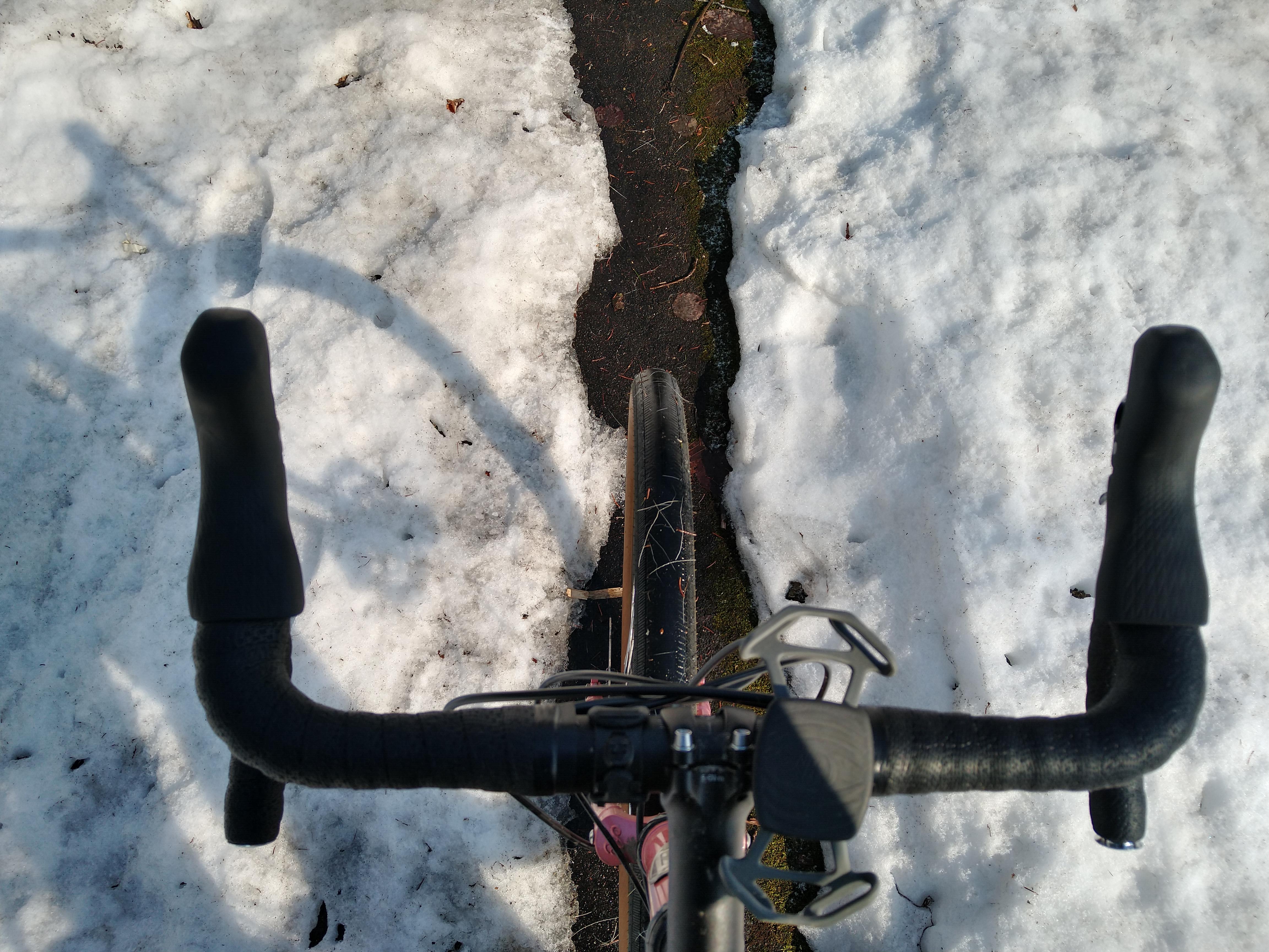 Looking down from a bike saddle while threading through a narrow strip of path with snow on either side.