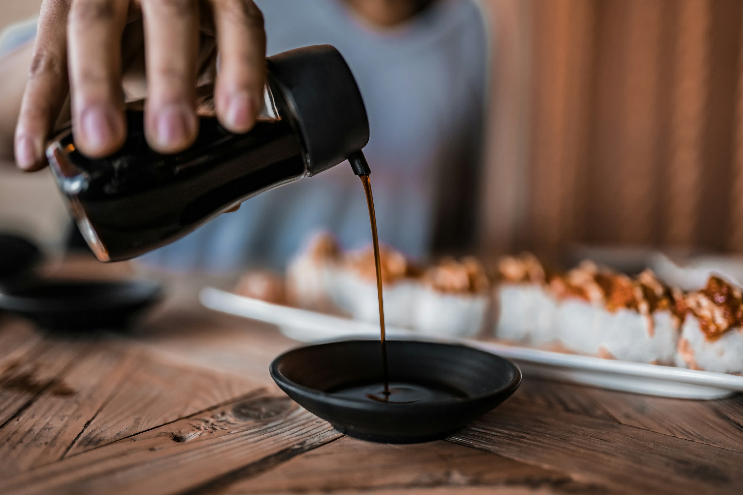 A hand holds a soy sauce dispenser and pours some into a small dish. There is a plate of sushi out of focus in the background.