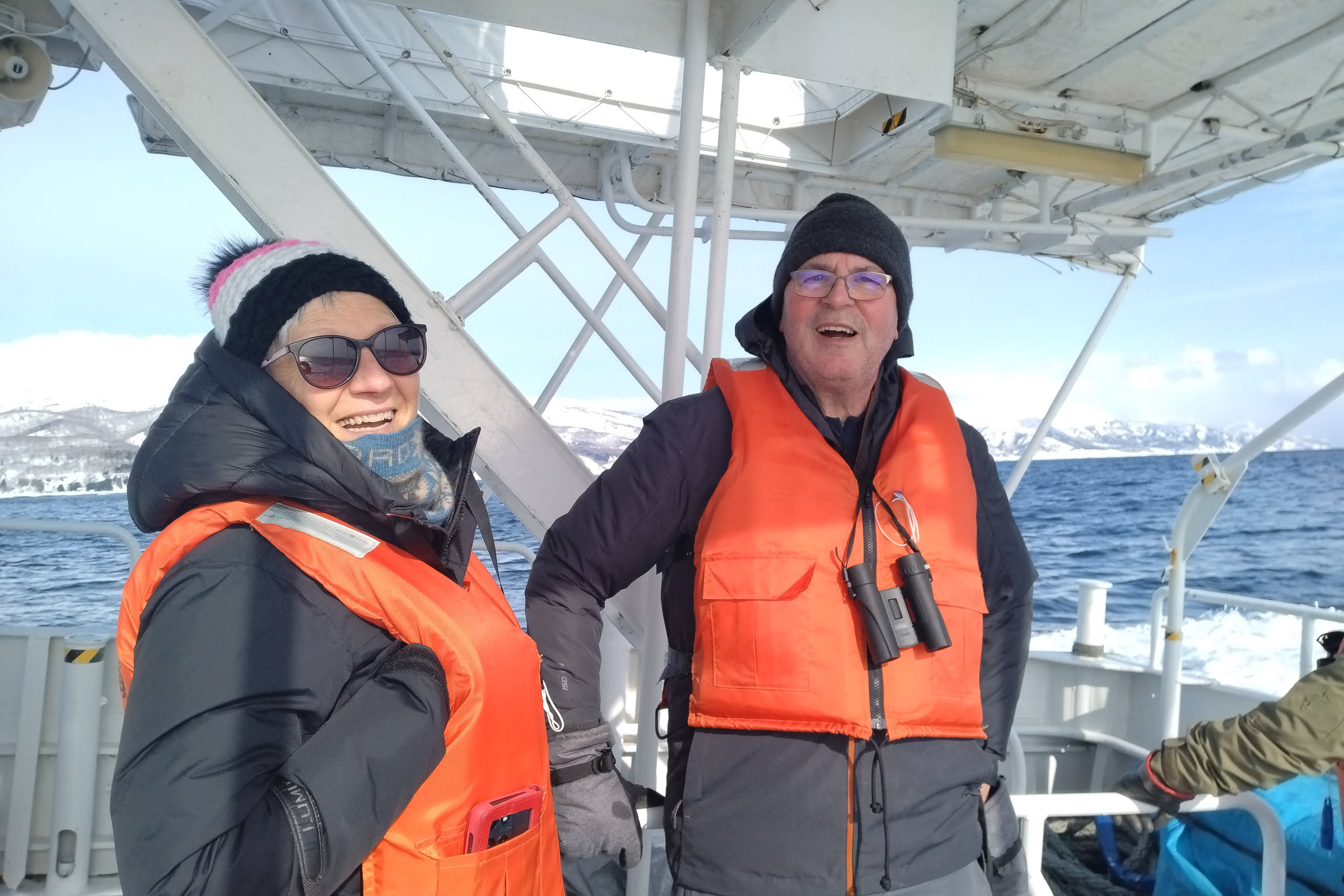 British couple enjoying a winter cruise along the Rausu Coast, smiling at camera while wearing life vests and warm winter clothing.