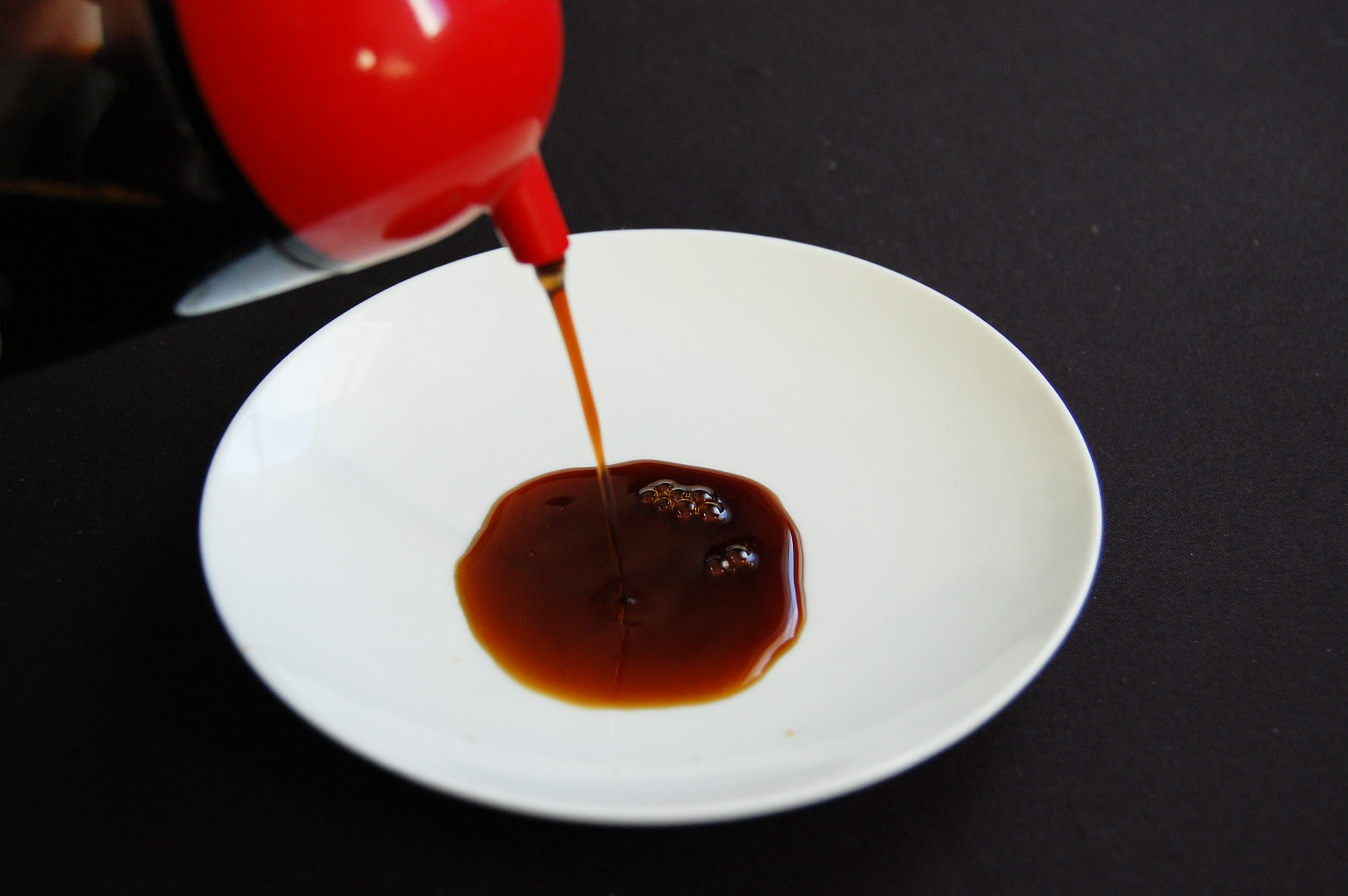 A small dish, into which a soy sauce dispenser is pouring a small amount of soy sauce.