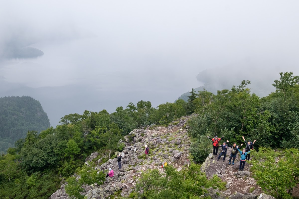 A group of hikers wave to the camera near the summit of Mt. Hakunzan. Lake Shikaribetsu is seen peaking out of the fog way bellow them.