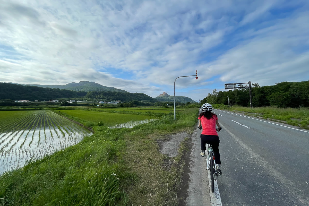 A cyclist rides past rice fields near Lake Toya with an active volcano in the background.