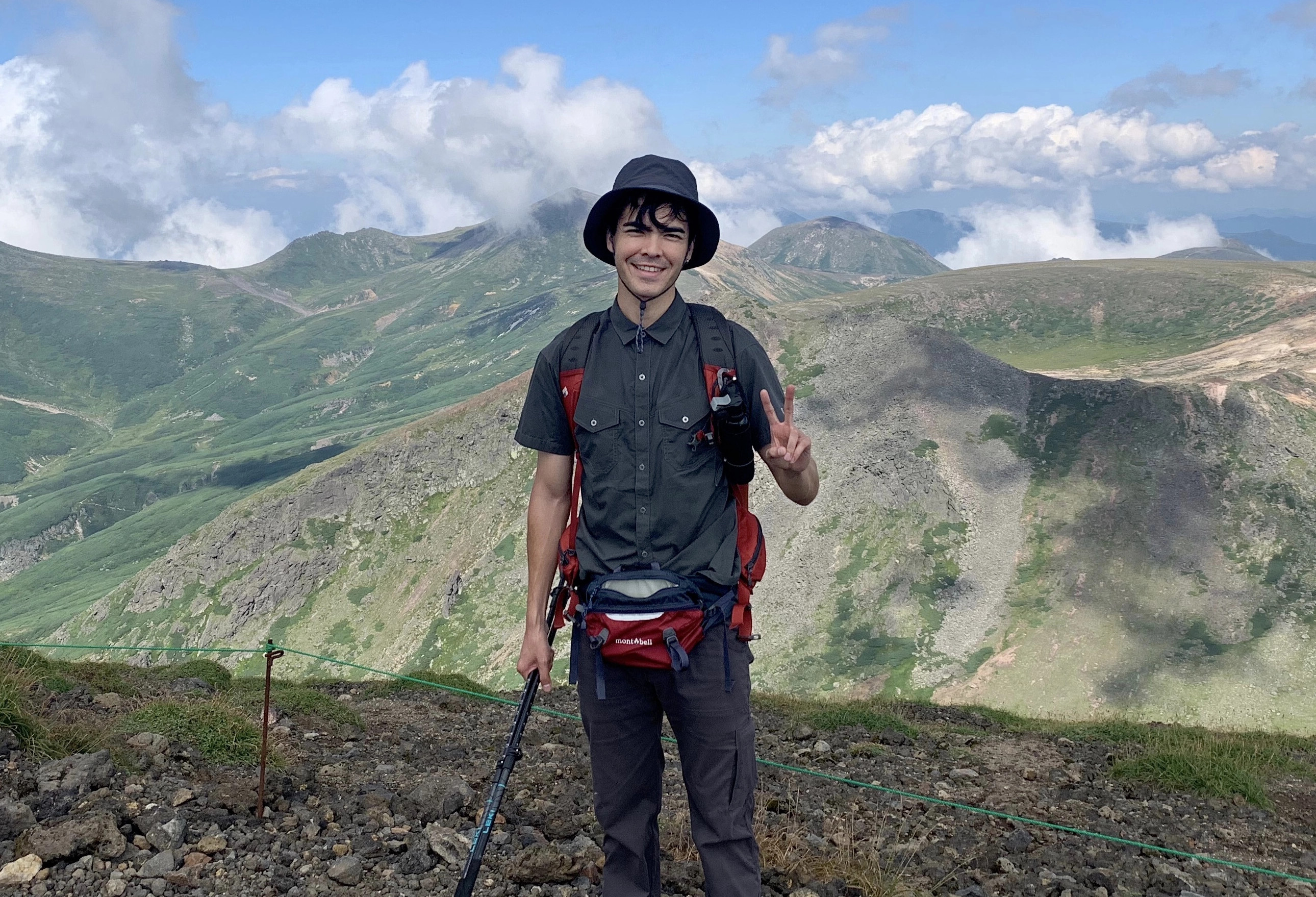 A hiker flashes a peace sign while posing on the slopes of Mt Asahidake. The Daisetsuzan mountains can be seen in the background.