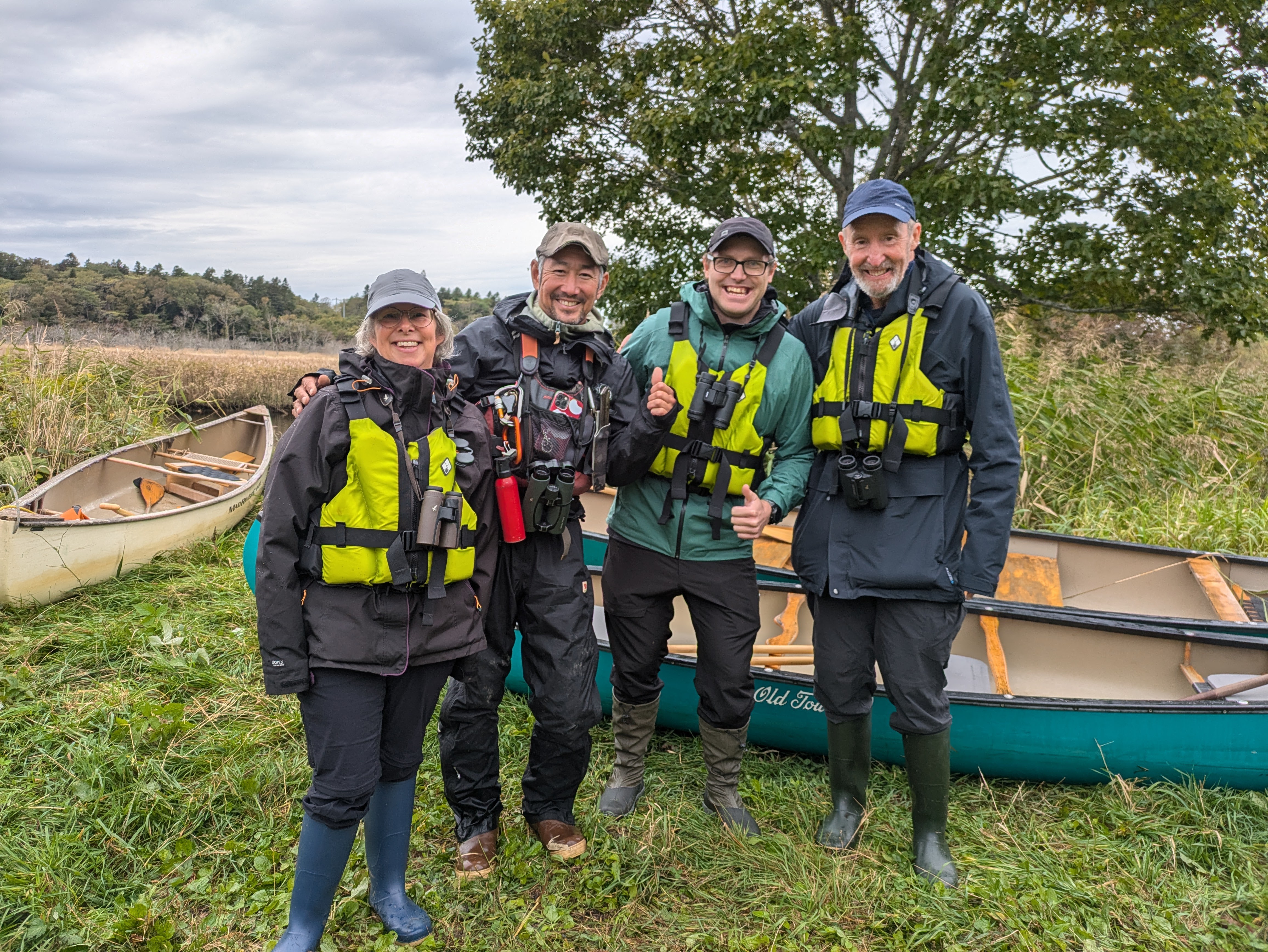 Four people stand in front of a canoe on a grassy bank and smile at the camera. They are all wearing life jackets. It is an overcast day.