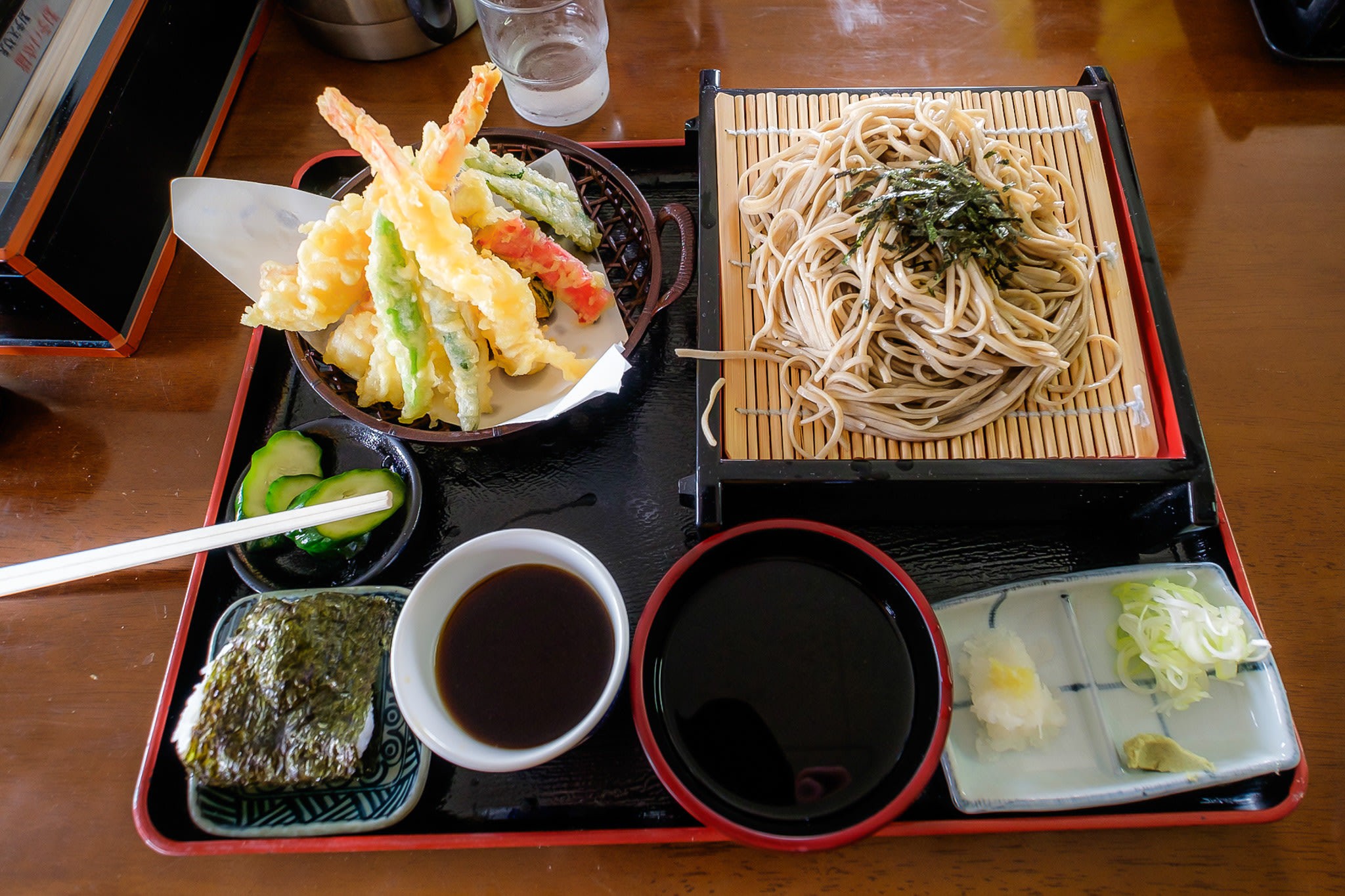 A Japanese "teishoku" style dish, with a small rice bowl, soba noodles, tempura, cucumber pickles and a dipping sauce for both noodles and tempura. There are toppings for the soba noodles on small plates.