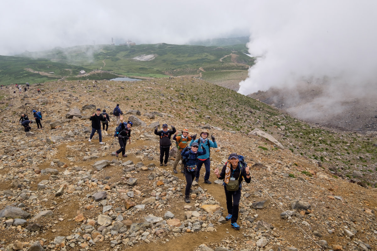 Hikers pose for a shot on the slope of Mt. Asahidake with steam from fumaroles in the background meeting the clouds.
