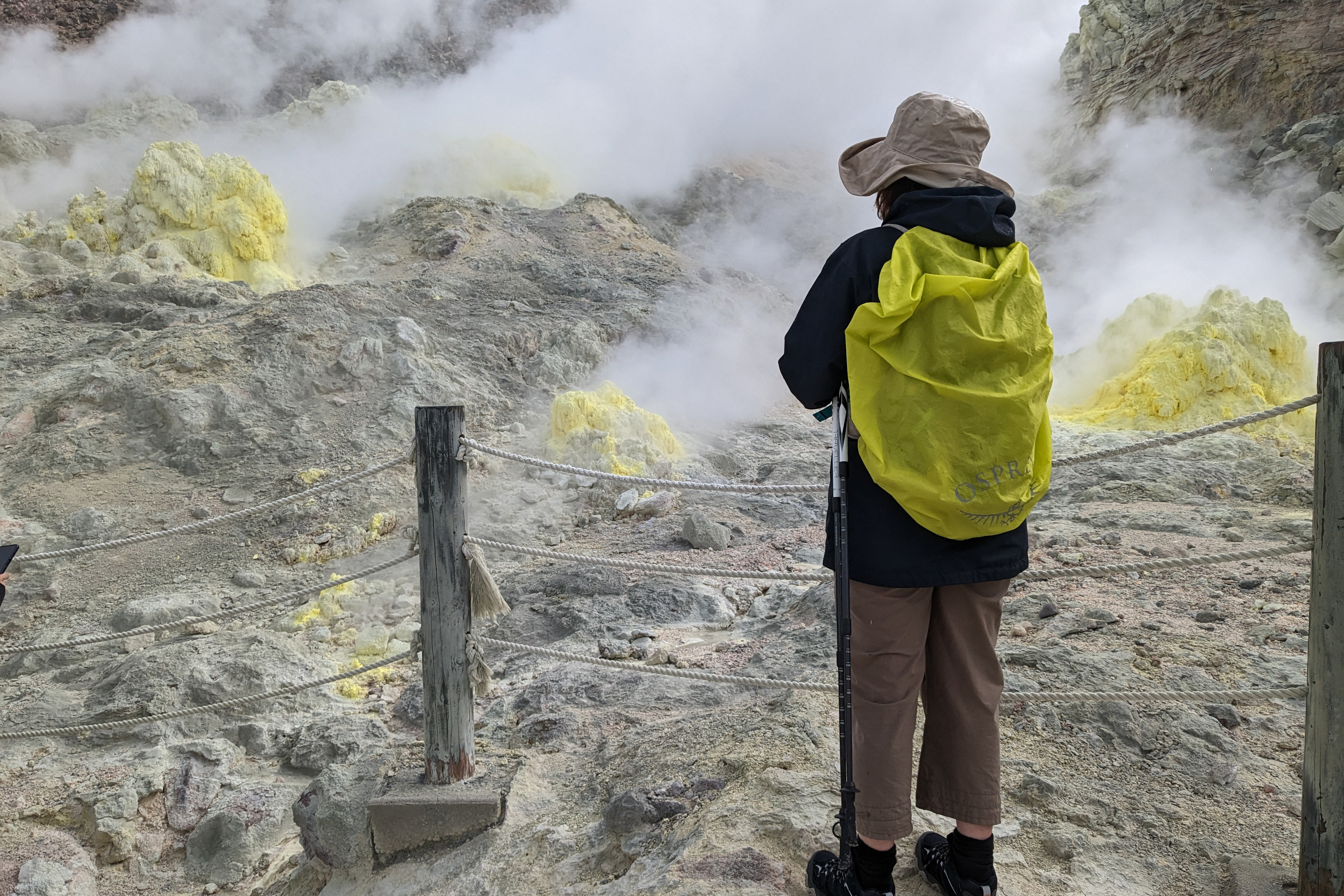 A tourist with a yellow pack cover stands in front of steaming volcanic vents that are coated in yellow sulpher