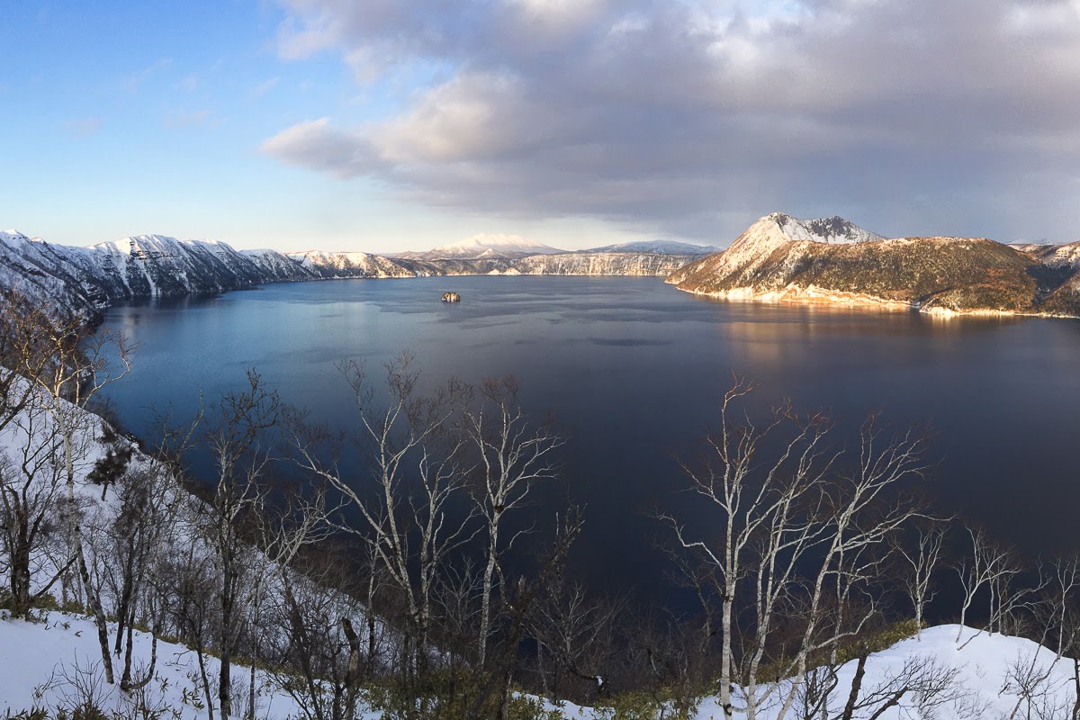 Mt Mashu, light by evening sun, is reflected in a still Lake Mashu. The ground is covered in early season snow.