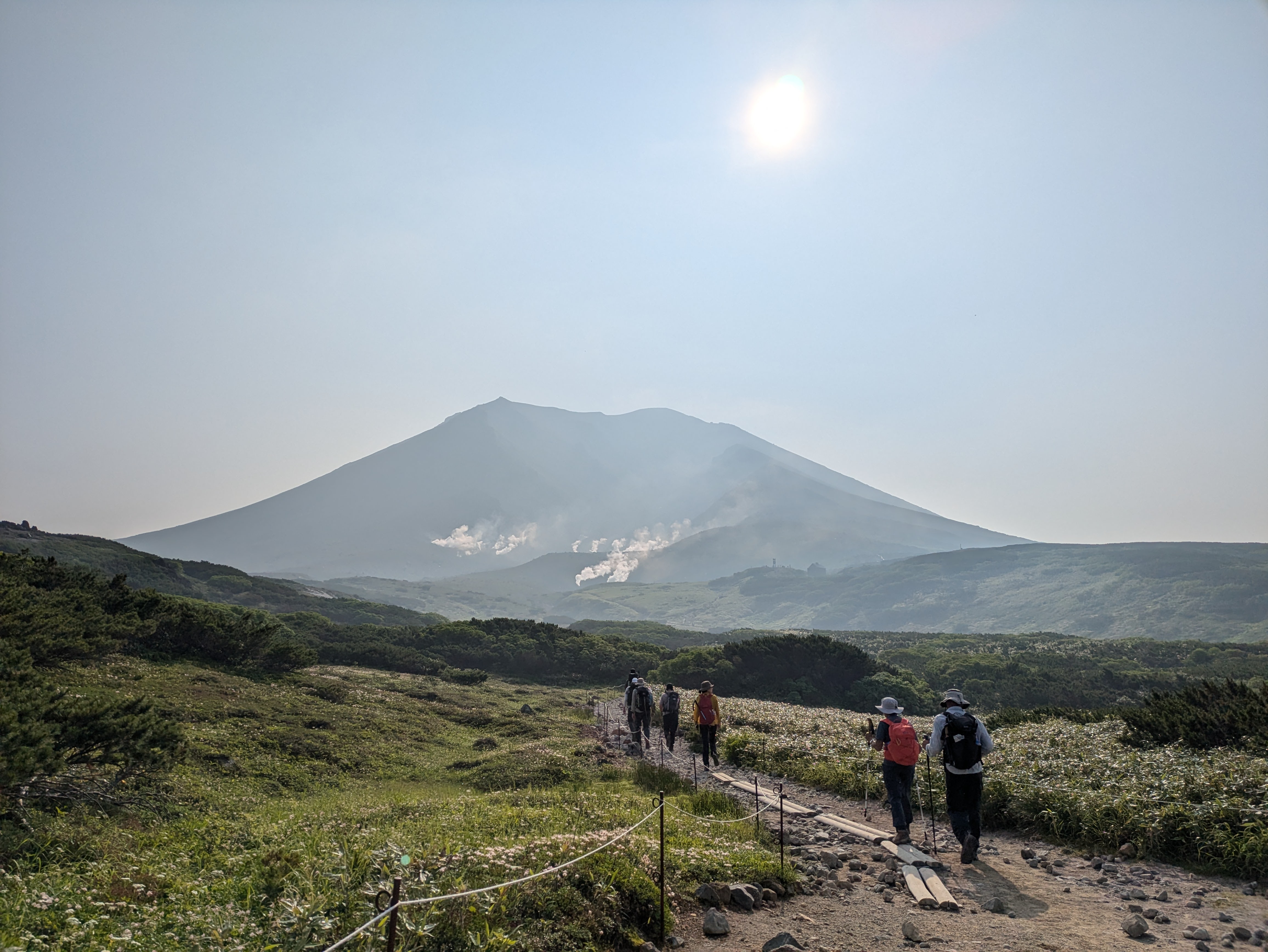 A line of hikers makes their way along a boardwalk towards the peak of Mt. Asahidake, the tallest mountain in Hokkaido. It is a very sunny day.