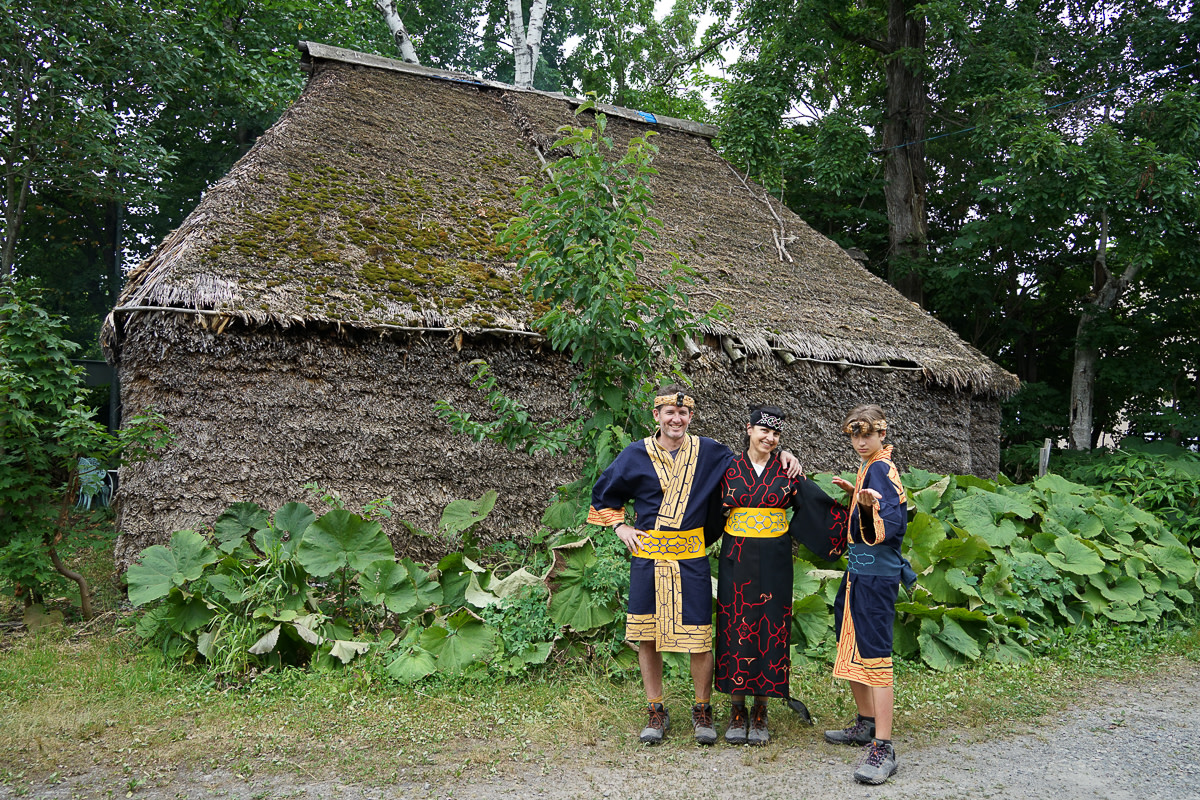 A family of travellers dressed in traditional Ainu clothing pose in front of a traditional Cise house