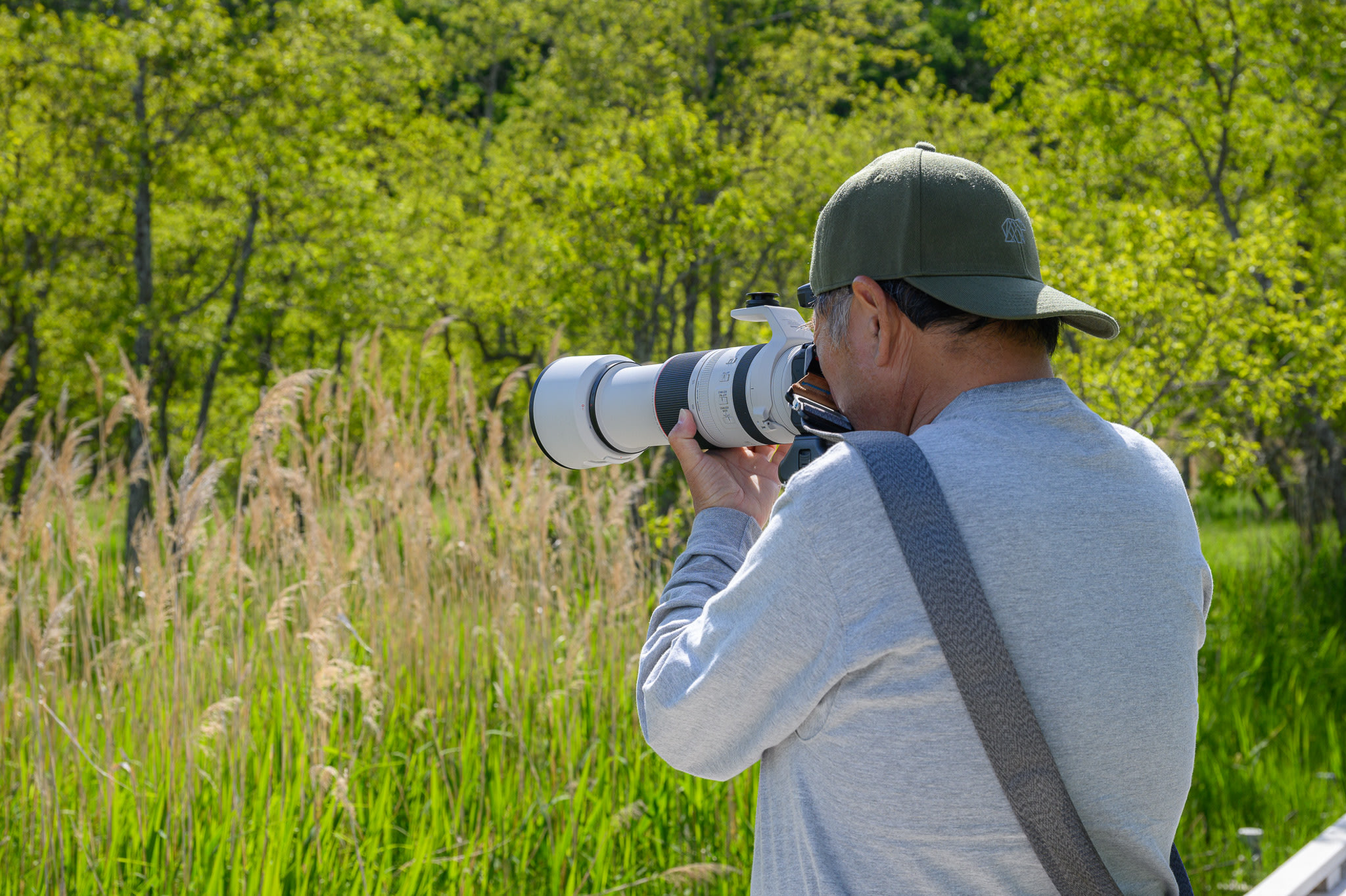 A man with his back to us holds a camera to his face. His camera is equipped with a long lens, which he is using to zoom in on a subject far away.