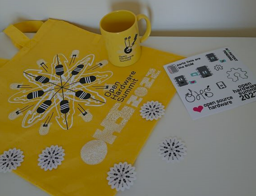the image contains some items from the 2022 open hardware summit goodie bag. the is a yellow bag laying on a table with the 2022 OHS logo on it, on top is a yellow mug featuring the logo and sprinkled on the table are stickers that say "OHS2022" and "<3 open source"