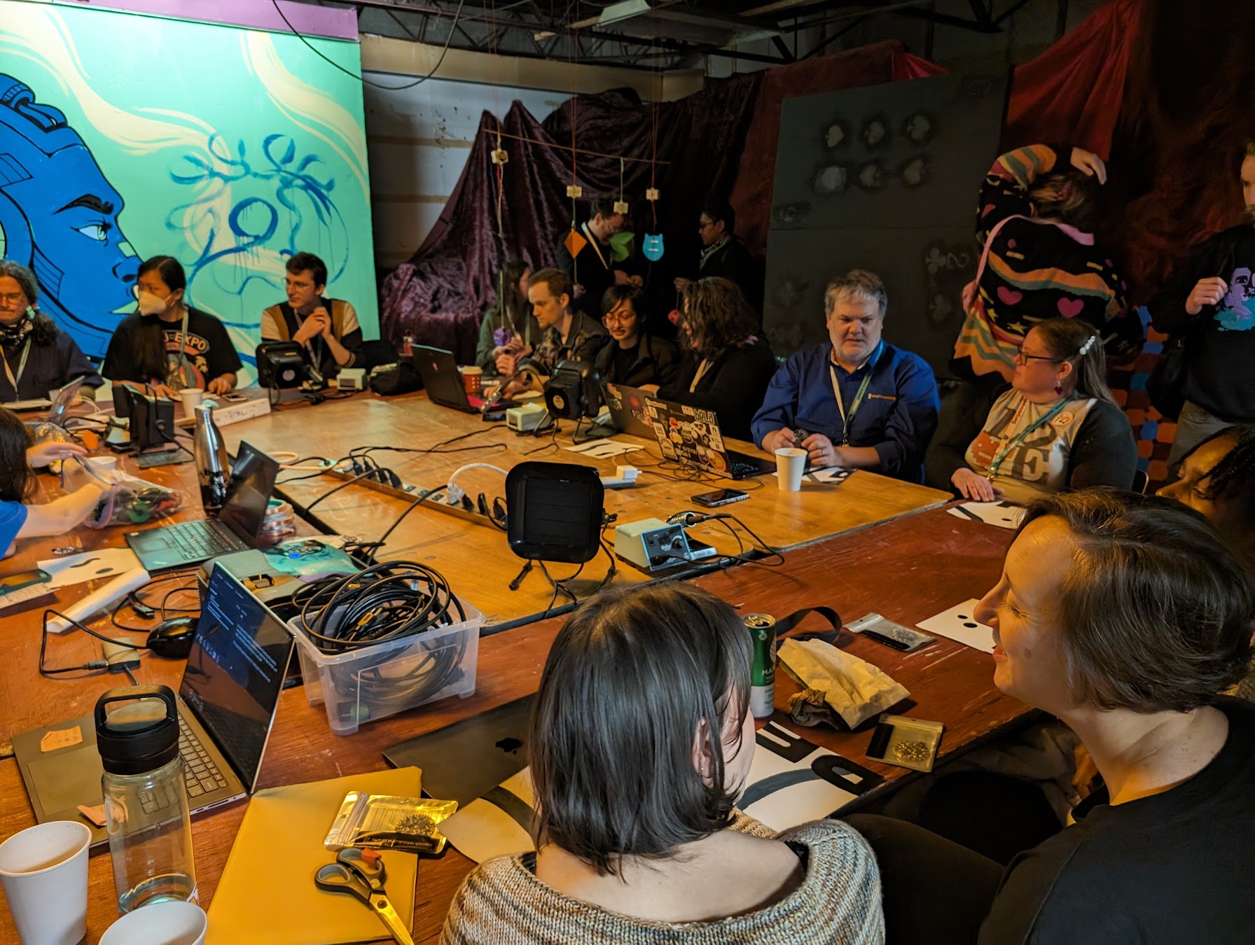 Open Hardware Summit attendees gather around a table of electronics for a session.