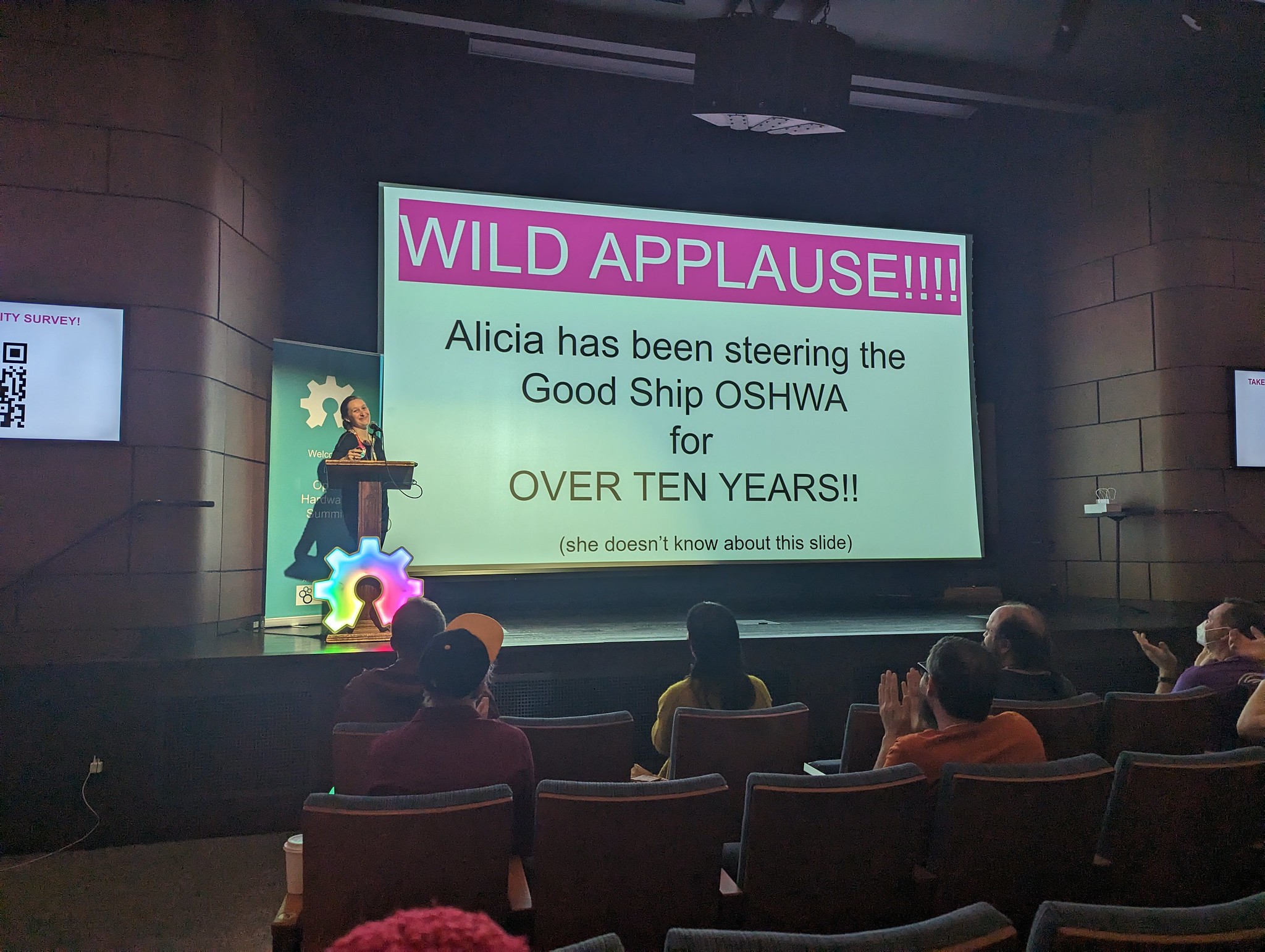 A photo of Alicia Gibb Seidle speaking at OHS 2023 with a surprise slide in her deck that says "Wild applause! Alicia has been steering the Good Ship OSHWA for over ten years! (she doesn't know about this slide)"