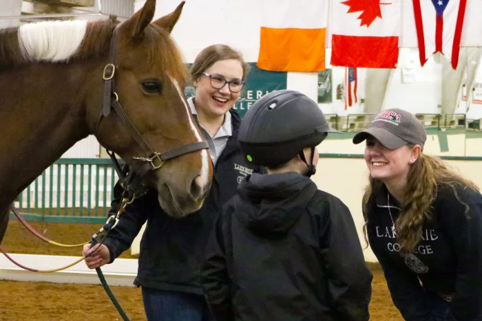 group of riding students smiling in a circle near a horse 