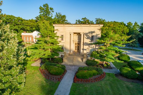 Cream, square block building with columns with gold front doors. Surrounded by grass and trees.