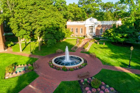 Arial shot of brick building with white columns and a fountain surrounded by a brick pathway and grass.