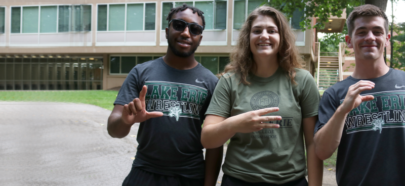 3 students in LEC tshirts outside the dorms spelling out L, E, C with their one hand on each of them.
