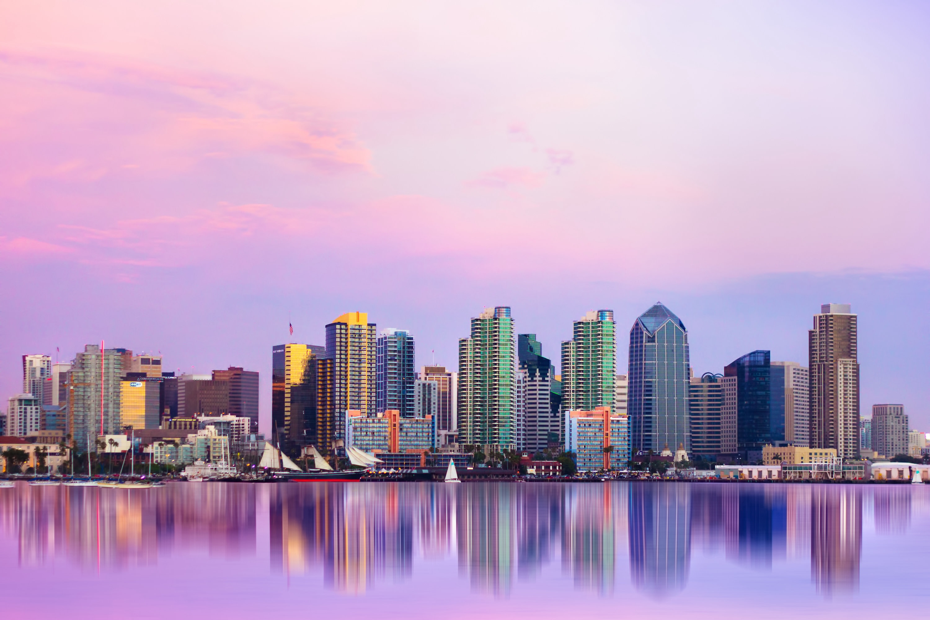 Photo of the skyline of San Diego with a purple and pink sunlit sky behind it that is also reflected in the water of the San Diego bay in front of it