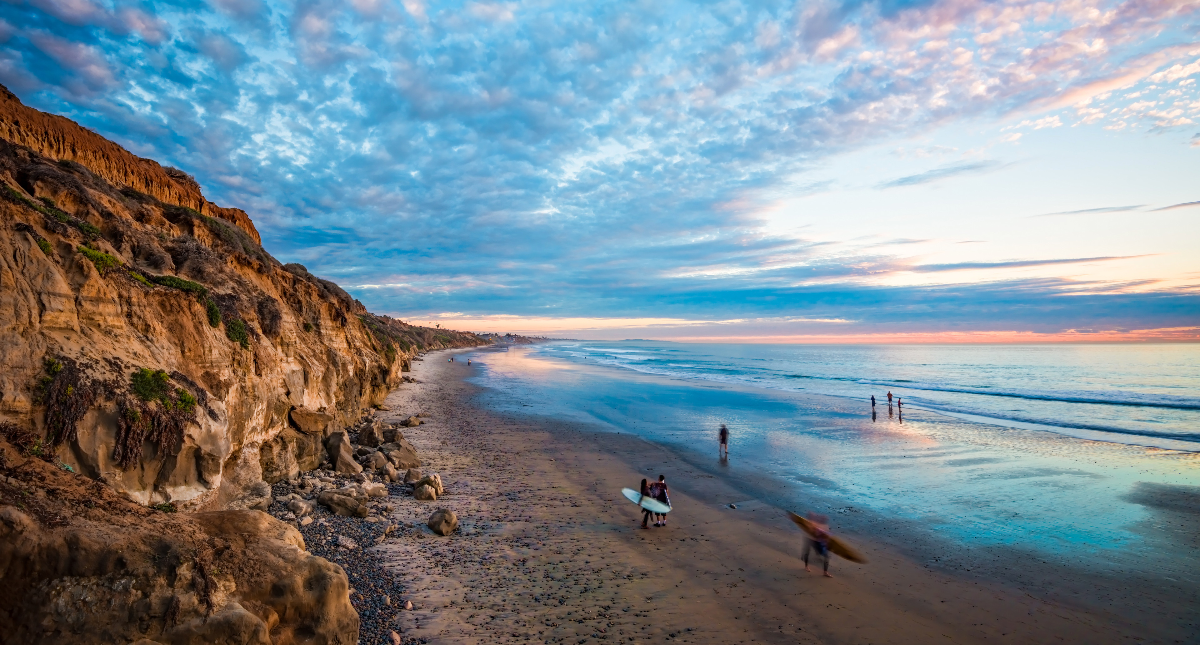 Photo of the beach and sandy cliffs in Carlsbad with surfers walking along the sand in the foreground and a blue, peach, and orange sunset in the background