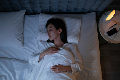 A woman in a white bed sleeping with a dimly lit reading lamp.