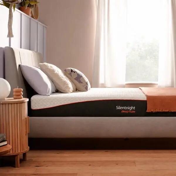 Silentnight mattress on a beige upholstered bed frame with orange throw, white pillows, and sheer curtains in background.