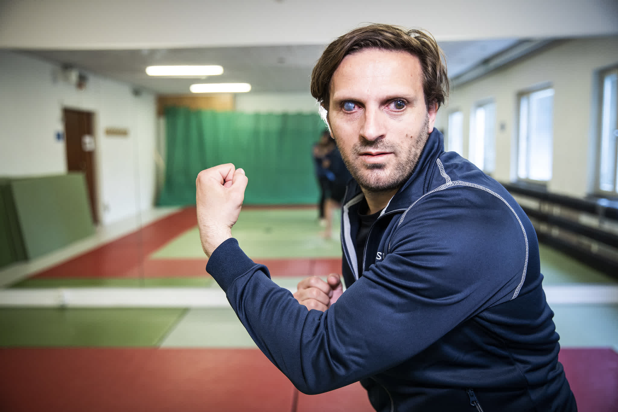 En man kollar in i kameran och har händerna i sparringposition. Han har en mörkblå, långärmad tröja, brunt hår och i bakgrunden syns en träningshall med judomatta.