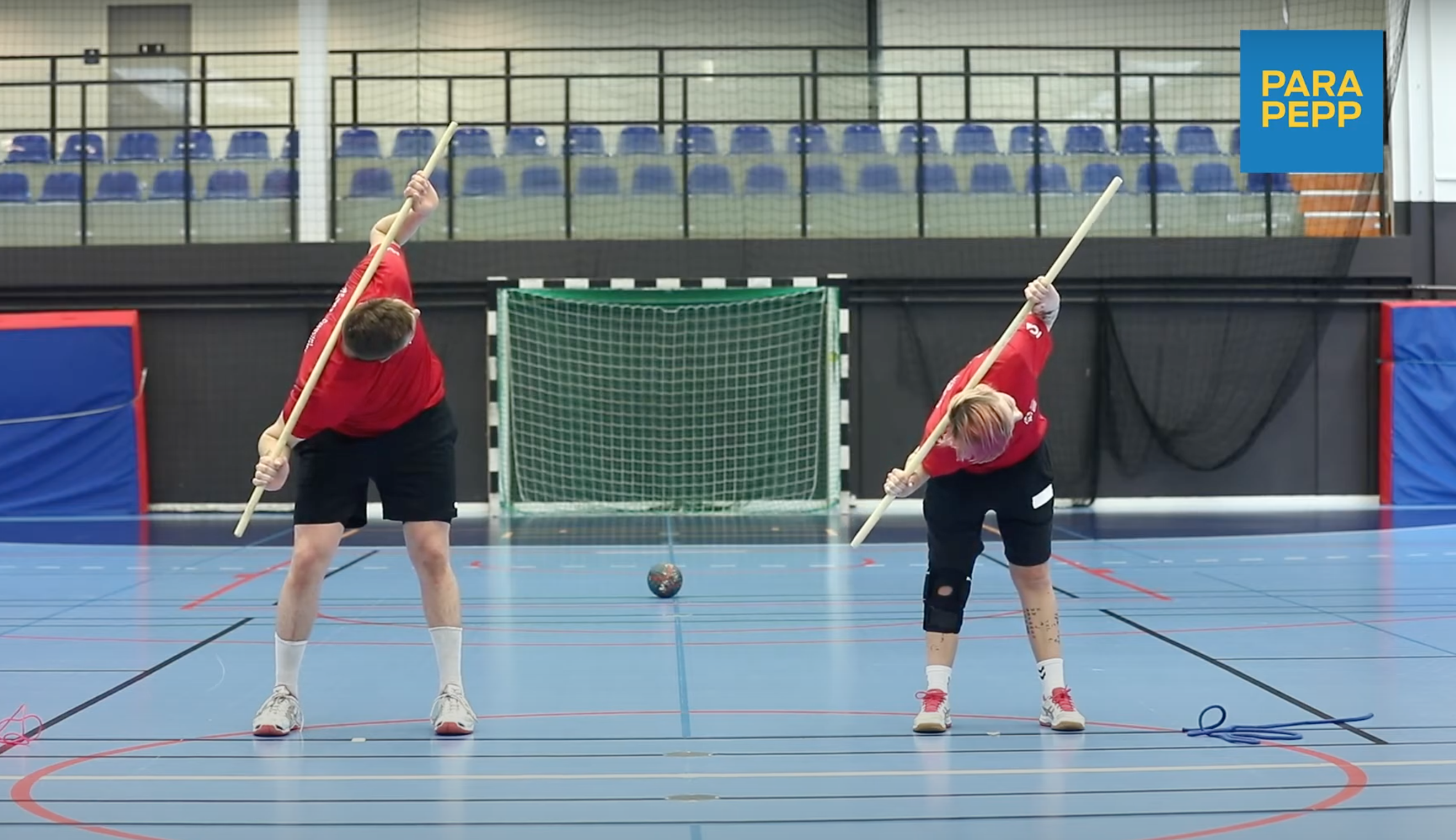 En man och kvinna står framåtböjda med en lång pinne bakom huvudet på axlarna. De har vridit överkroppen snett uppåt, med händerna på pinnen. De är i en stor idrottssal. 