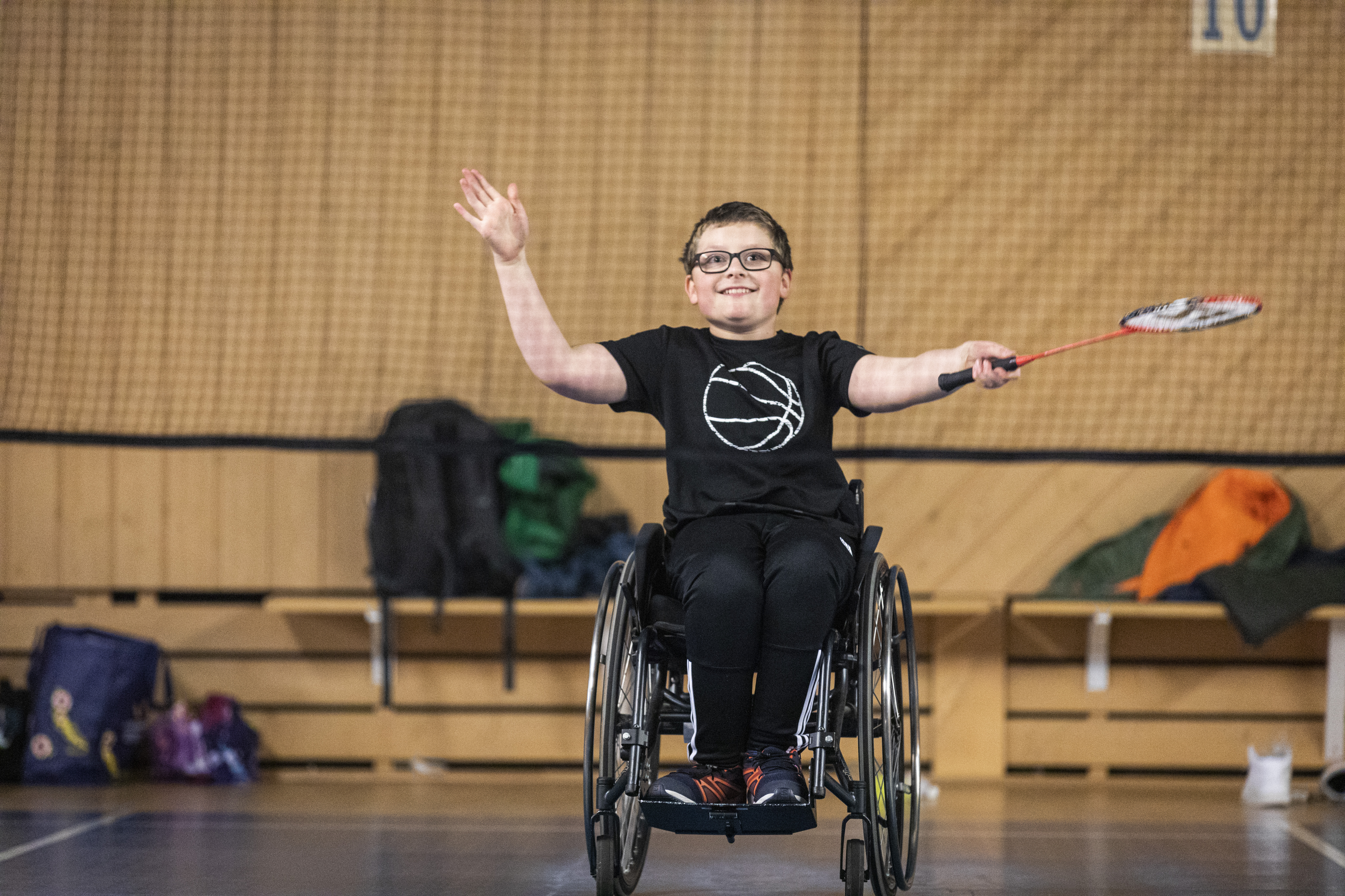 En pojke i rullstol sitter bakom badmintonnätet och väntar på att skjuta iväg badmintonfjädern. 