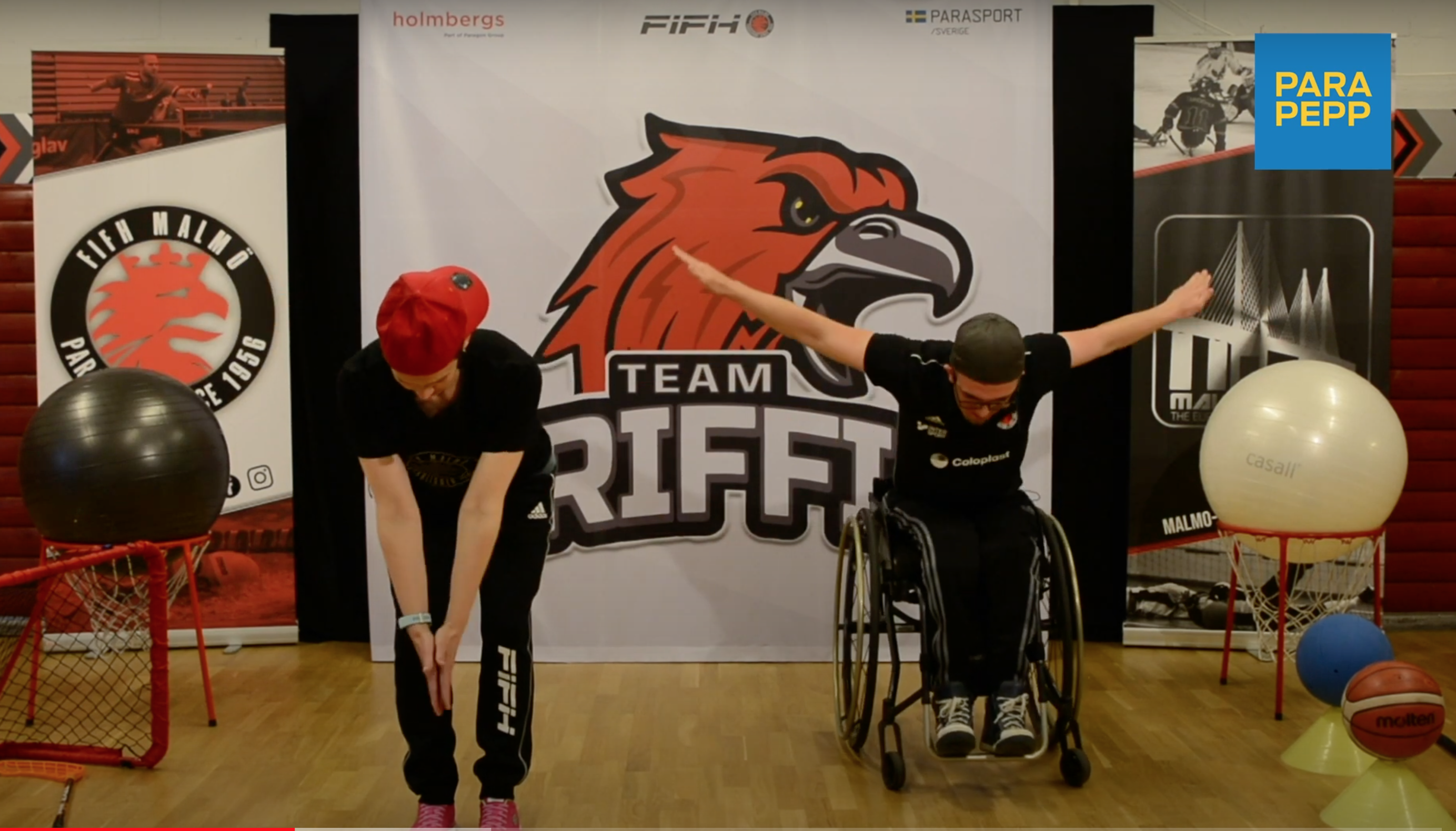 En kvinna som står upp och en man som sitter i rullstol. Kvinnan böjer sig framåt, har raka armar och handflatorna ihop och pekandes mot golvet. Mannen sitter lite framåtböjd, med armarna ut åt sidorna lite bakåt. 